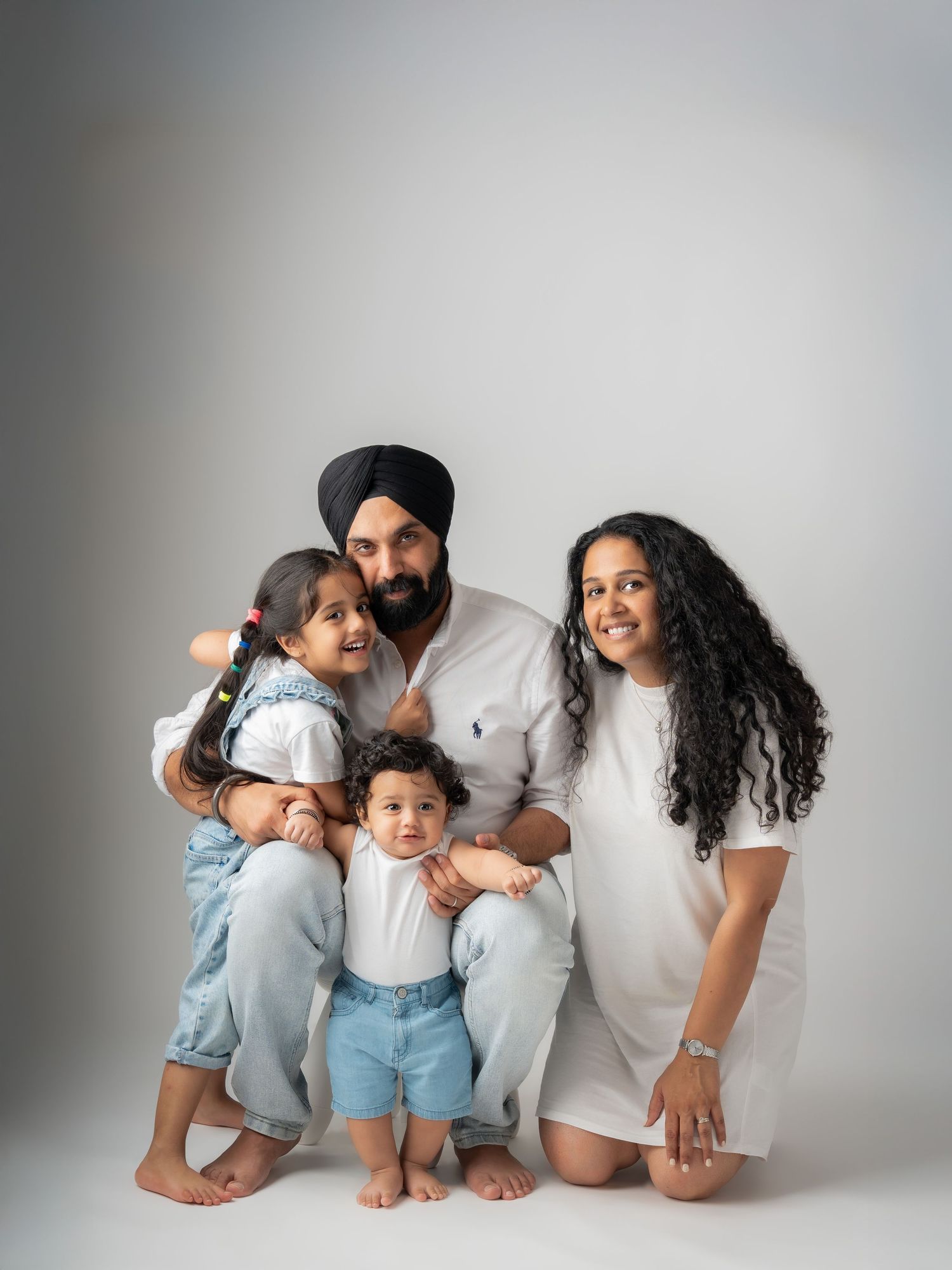Sikh family of four posing together in white outfits with baby and young daughter.