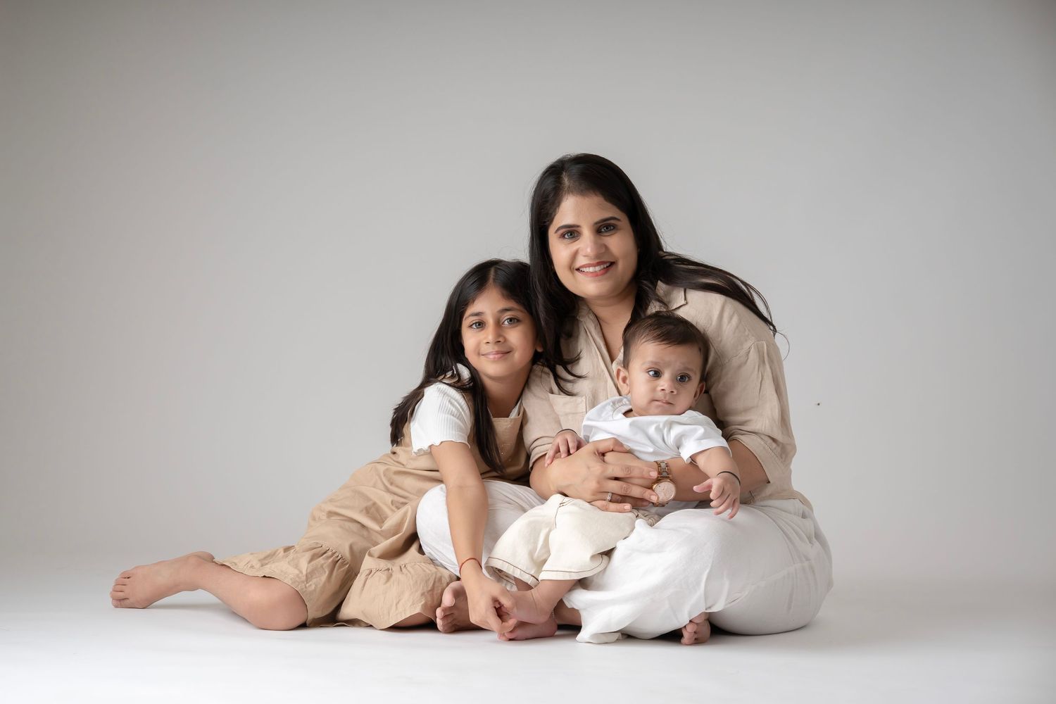 A smiling mother in beige sits cross-legged with her young daughter and baby in a bright studio portrait.