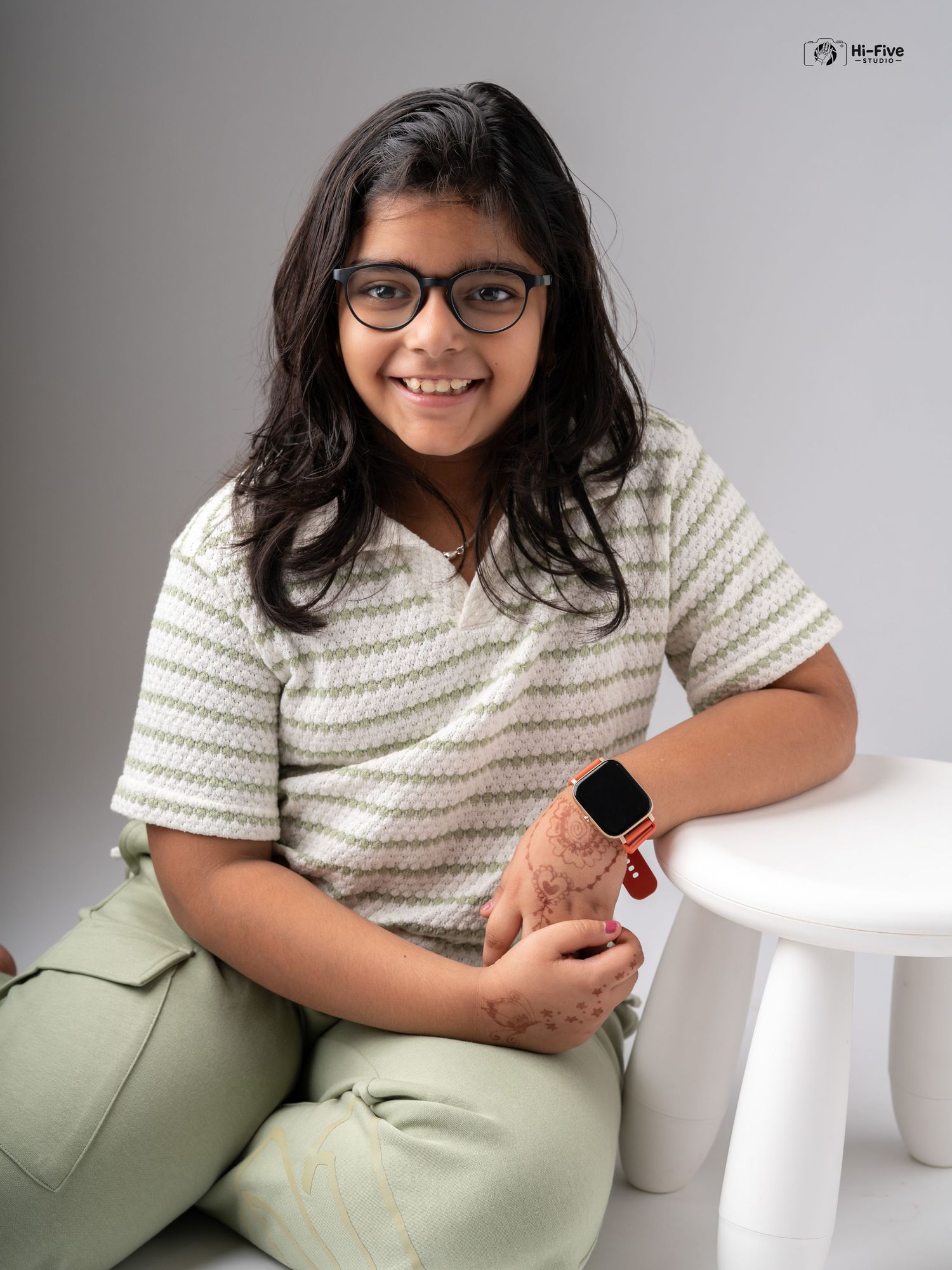 Smiling preteen girl with glasses wearing striped top and green pants with smartwatch on wrist.