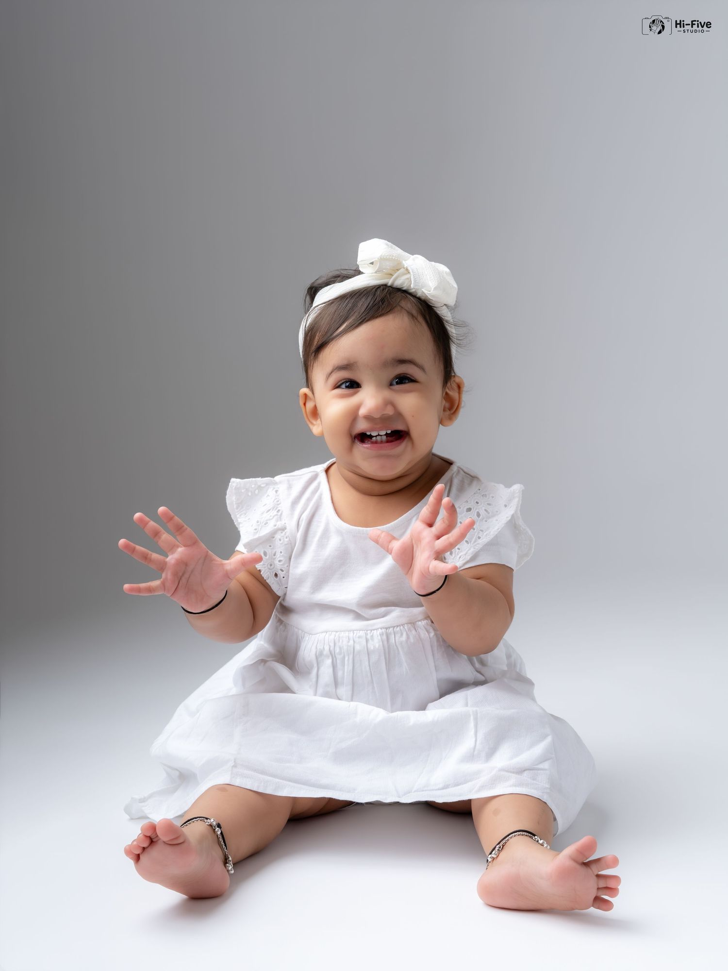Adorable baby girl in white dress and bow headband clapping hands and smiling in studio.