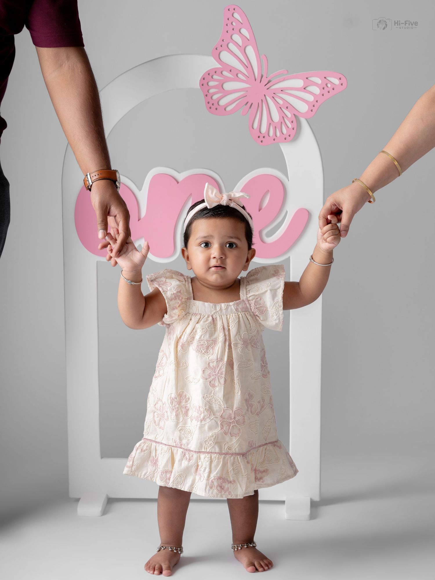 A baby girl in a floral dress stands holding hands at her first birthday photoshoot with a pink butterfly backdrop.