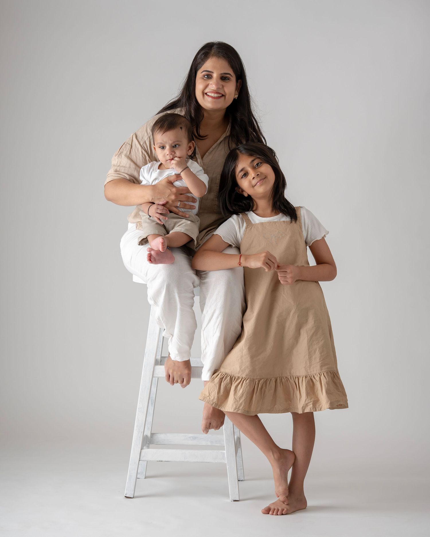 A happy mother perches on white stool holding infant while young daughter in tan pinafore dress leans against her.
