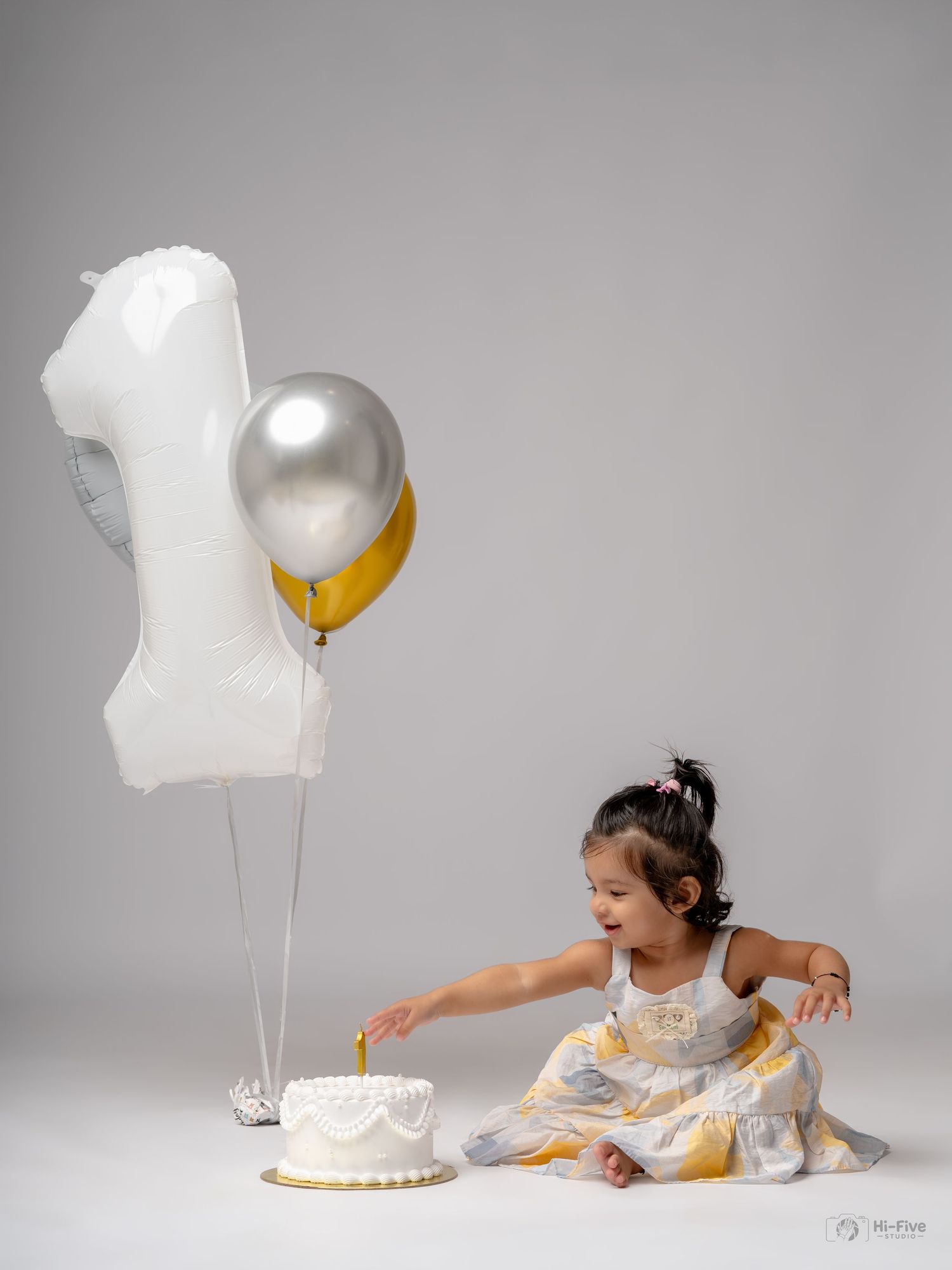 Baby girl in white dress touches first birthday cake beside number one balloon and silver gold balloons.