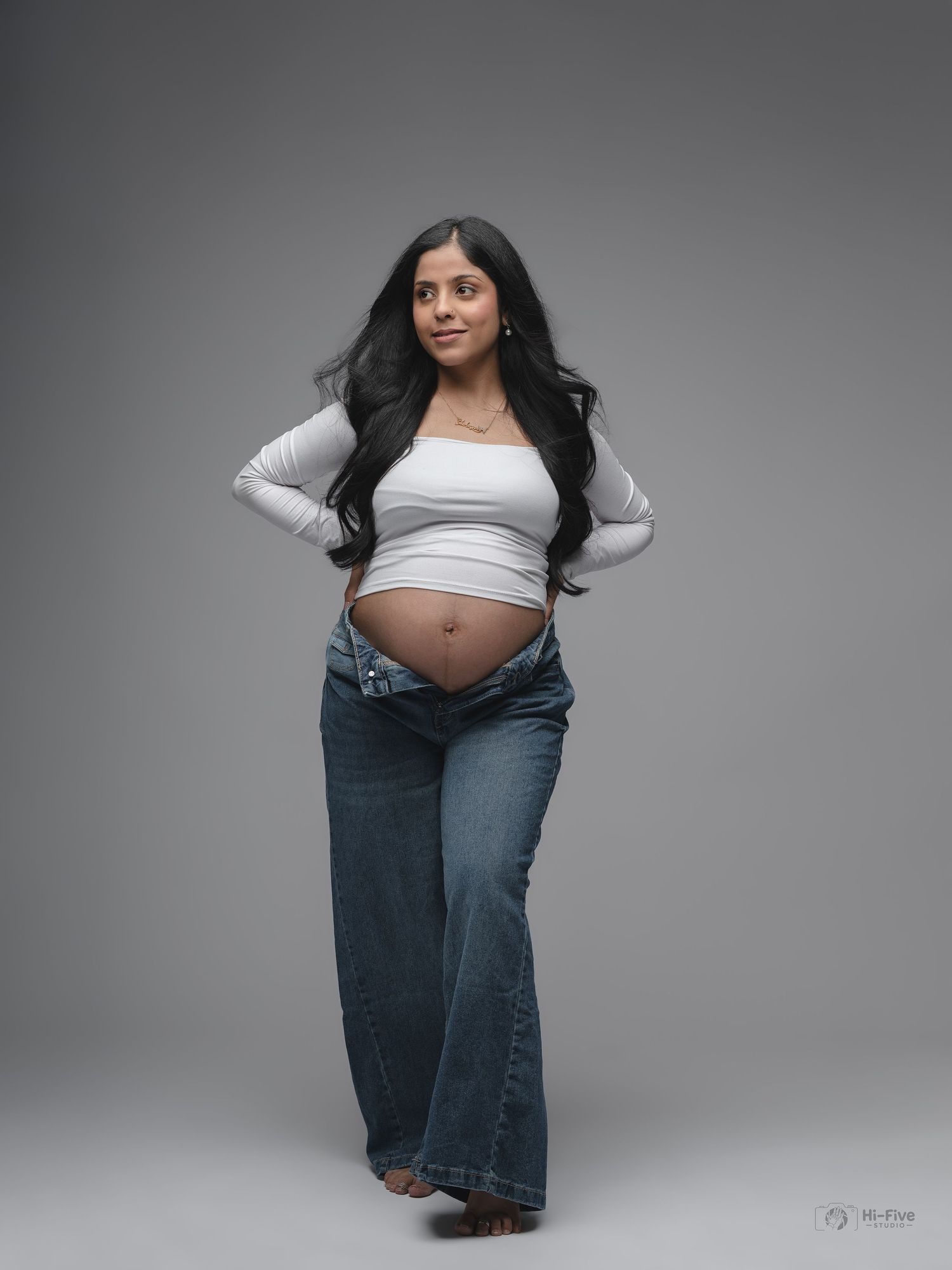 Pregnant woman in white crop top and jeans showing baby bump in professional maternity studio photo.