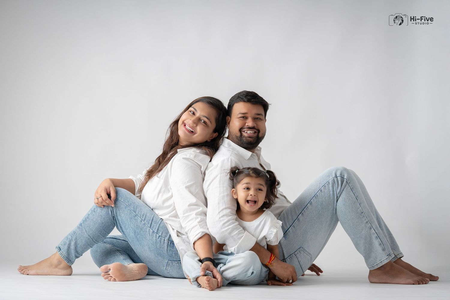 Happy family of three in white tops and jeans sitting back-to-back in a studio portrait.