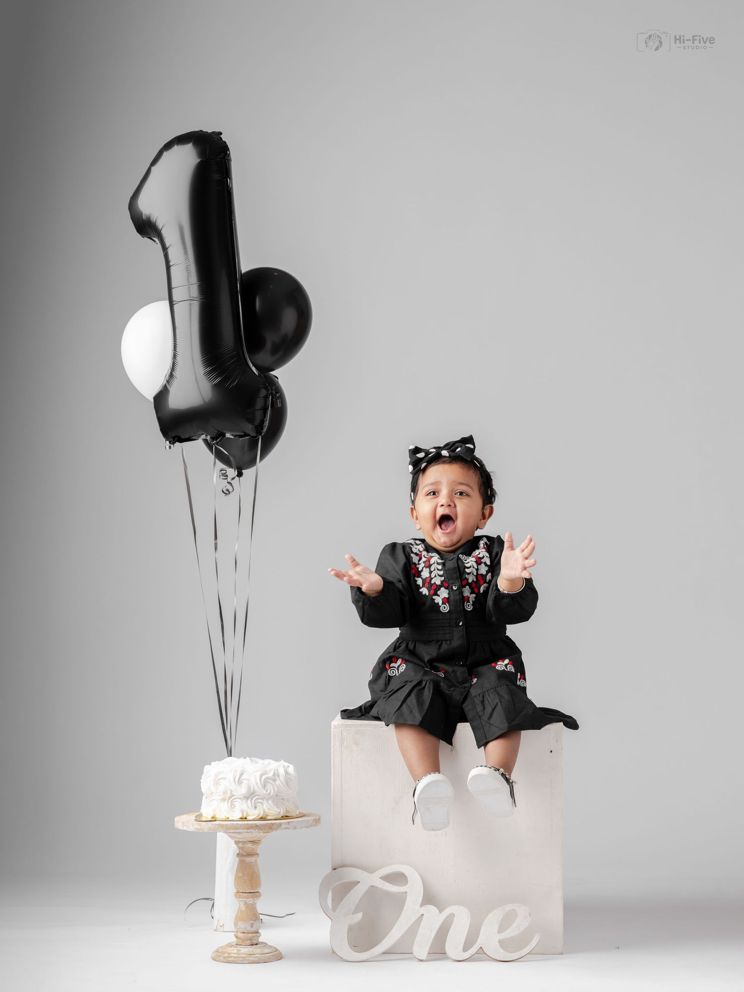 Baby girl in black dress celebrates first birthday with number one balloon and smash cake.