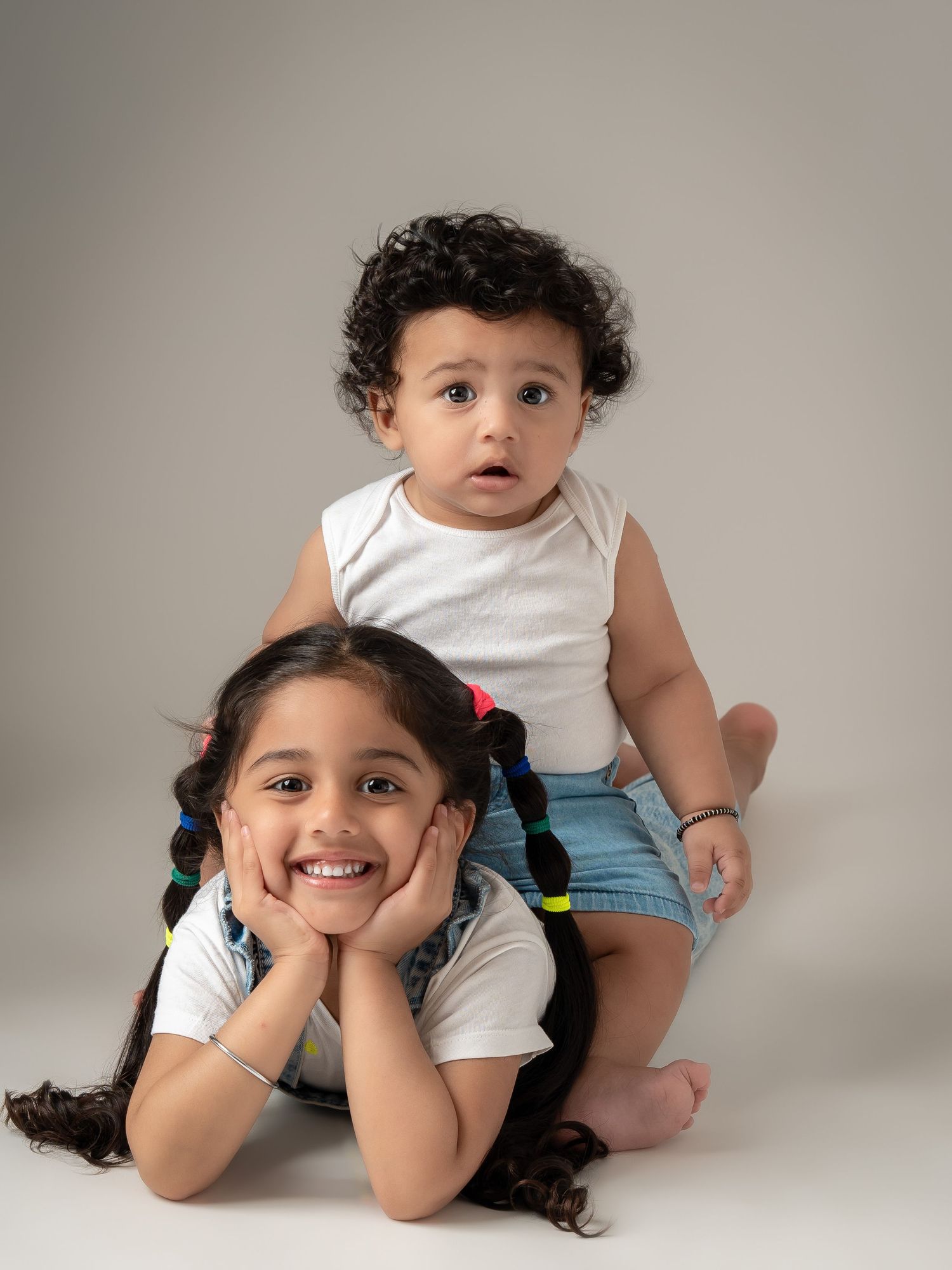 Adorable young girl with colorful braids posing with a curly-haired baby sibling in a studio portrait.
