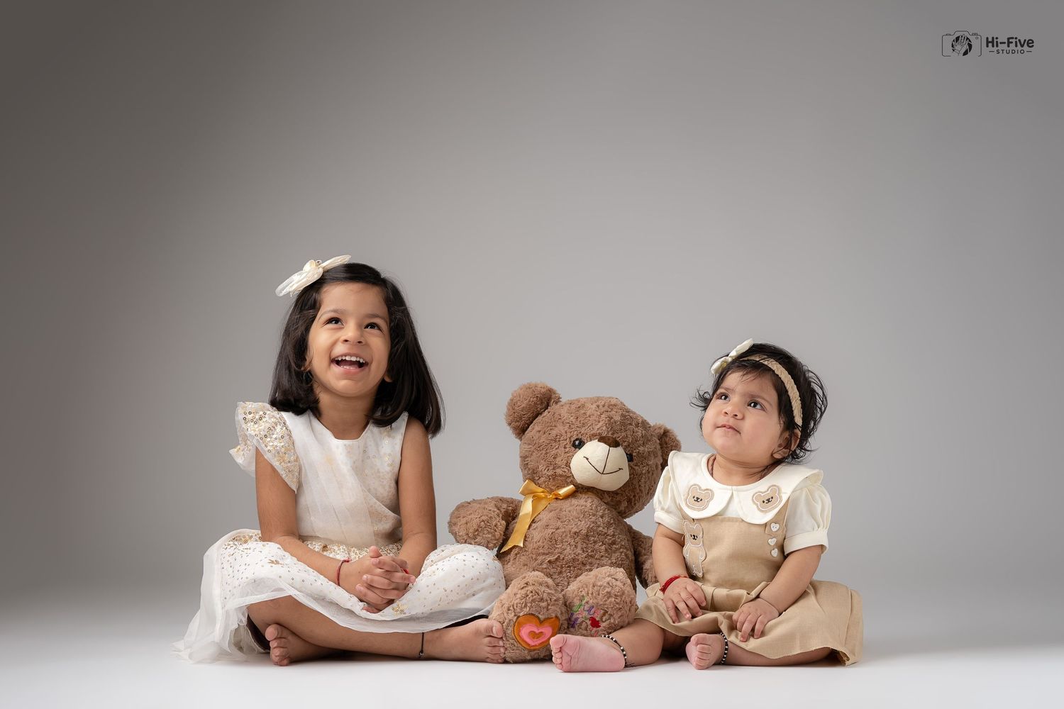 Two Indian sisters in white and cream dresses sit with a teddy bear in a professional studio photo.