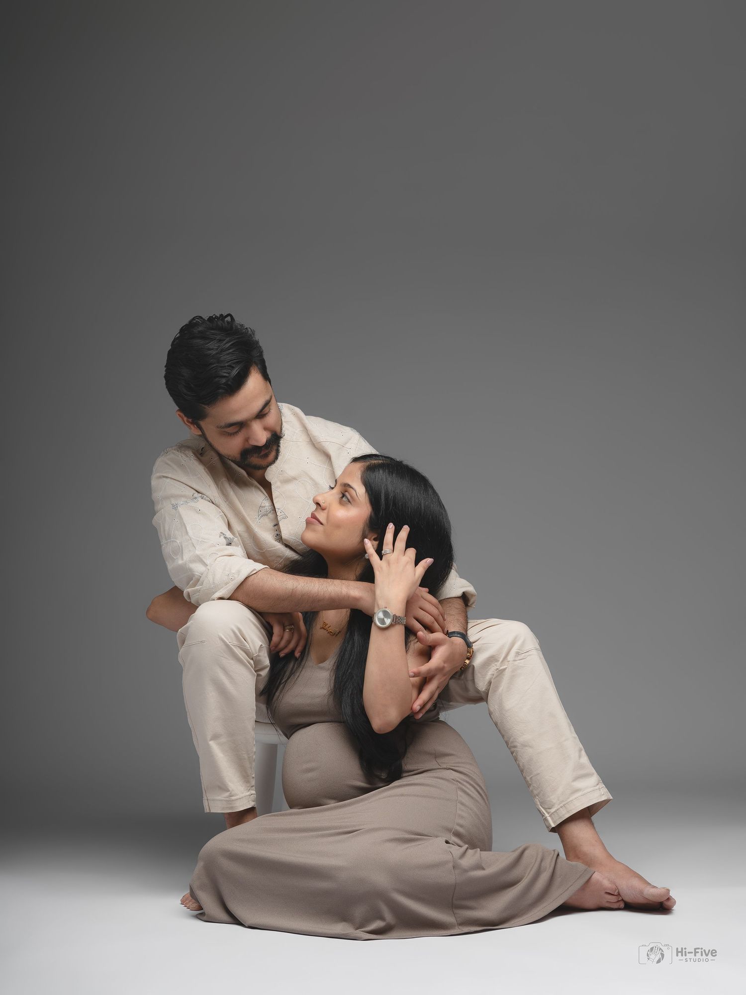 Couple maternity photoshoot with man embracing pregnant woman sitting on floor in beige outfits against gray background.