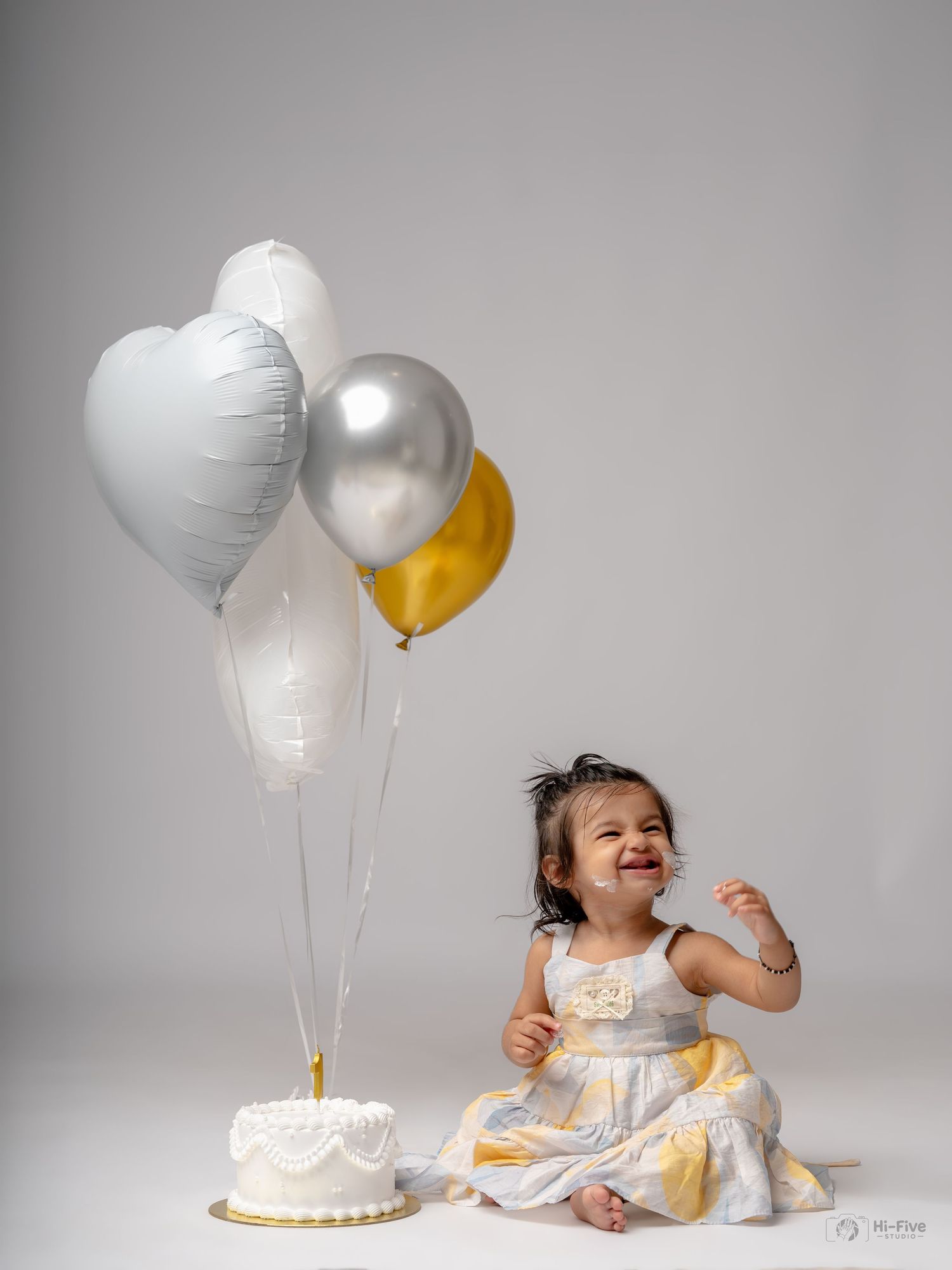 Smiling toddler girl in white yellow dress sits beside birthday cake with silver gold heart balloons in studio photo.