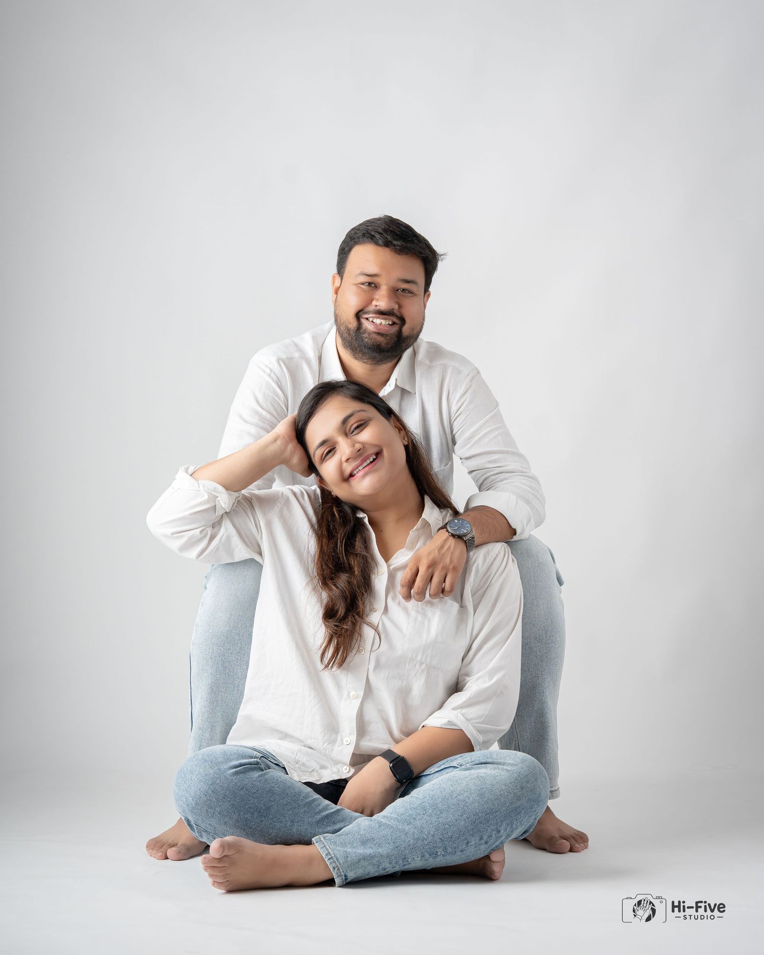Couple in matching white shirts and blue jeans posing together in a bright photography studio.