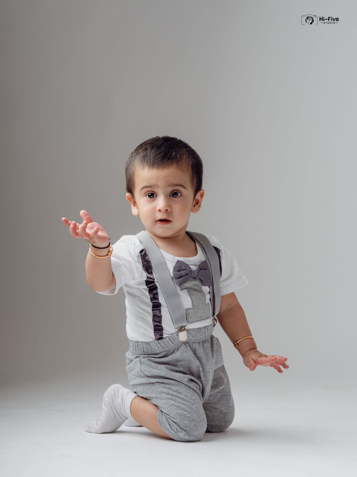 A cute toddler boy in a white shirt with suspenders and bow tie reaches out in a studio photo.
