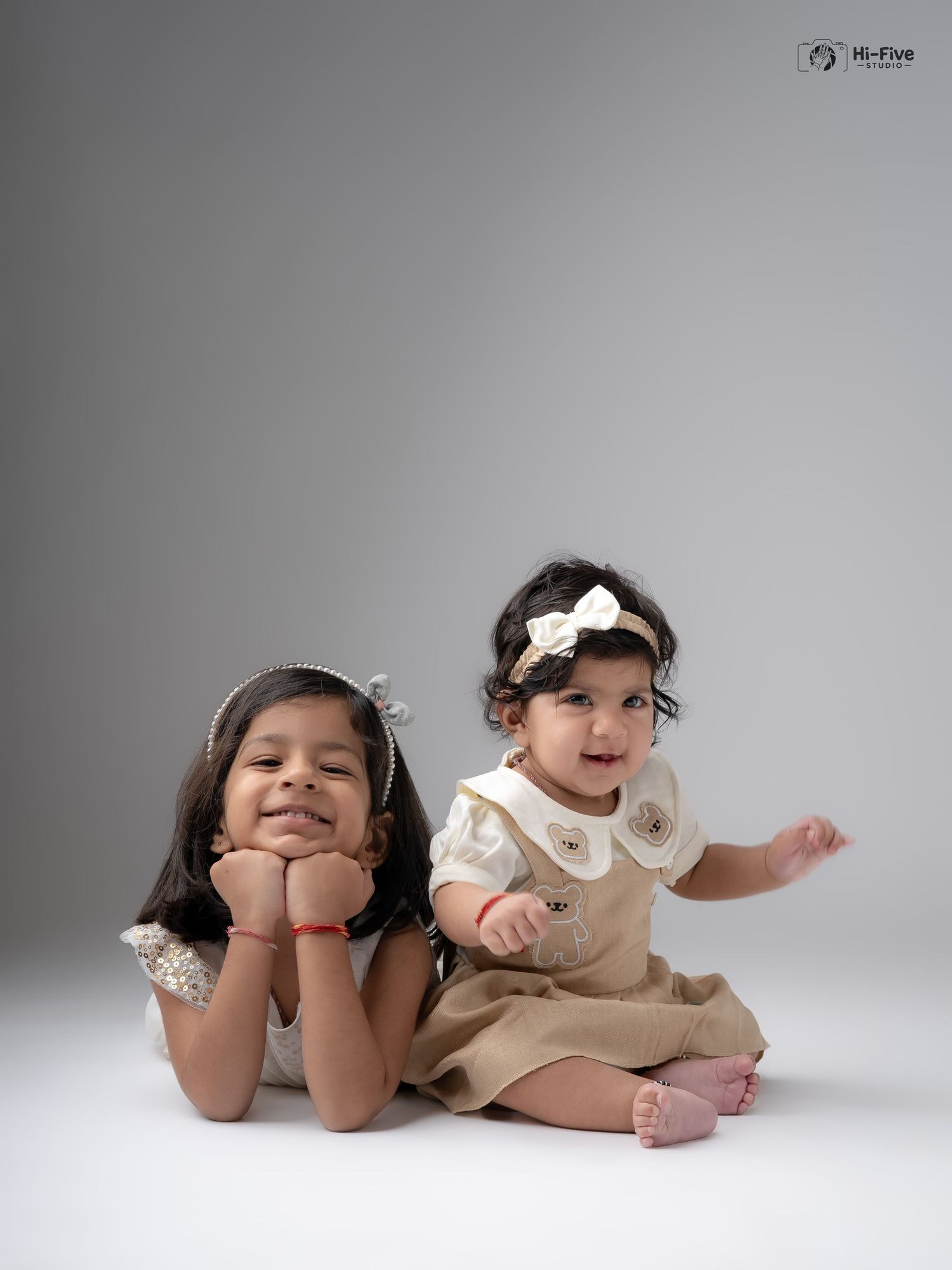 Two young Indian sisters in matching cream outfits pose playfully in a professional studio photography session.