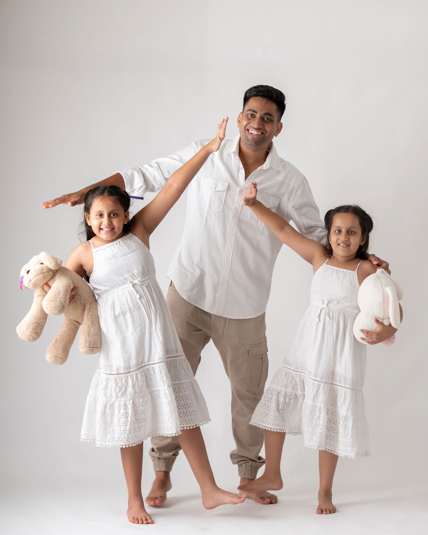A playful father and his two daughters strike fun poses in matching white outfits in a studio.