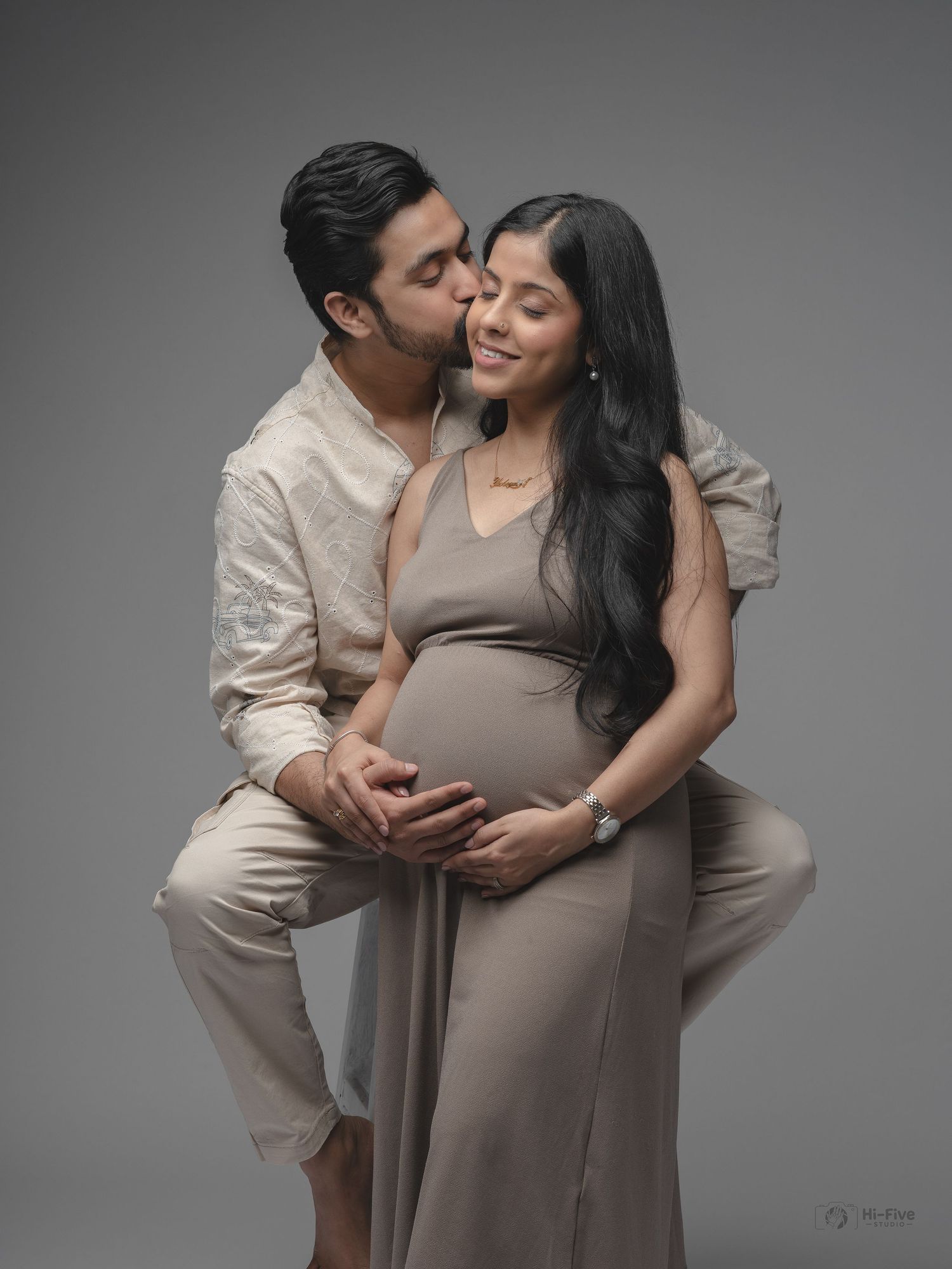 Man kissing pregnant partners cheek while embracing her baby bump in romantic maternity studio photo.