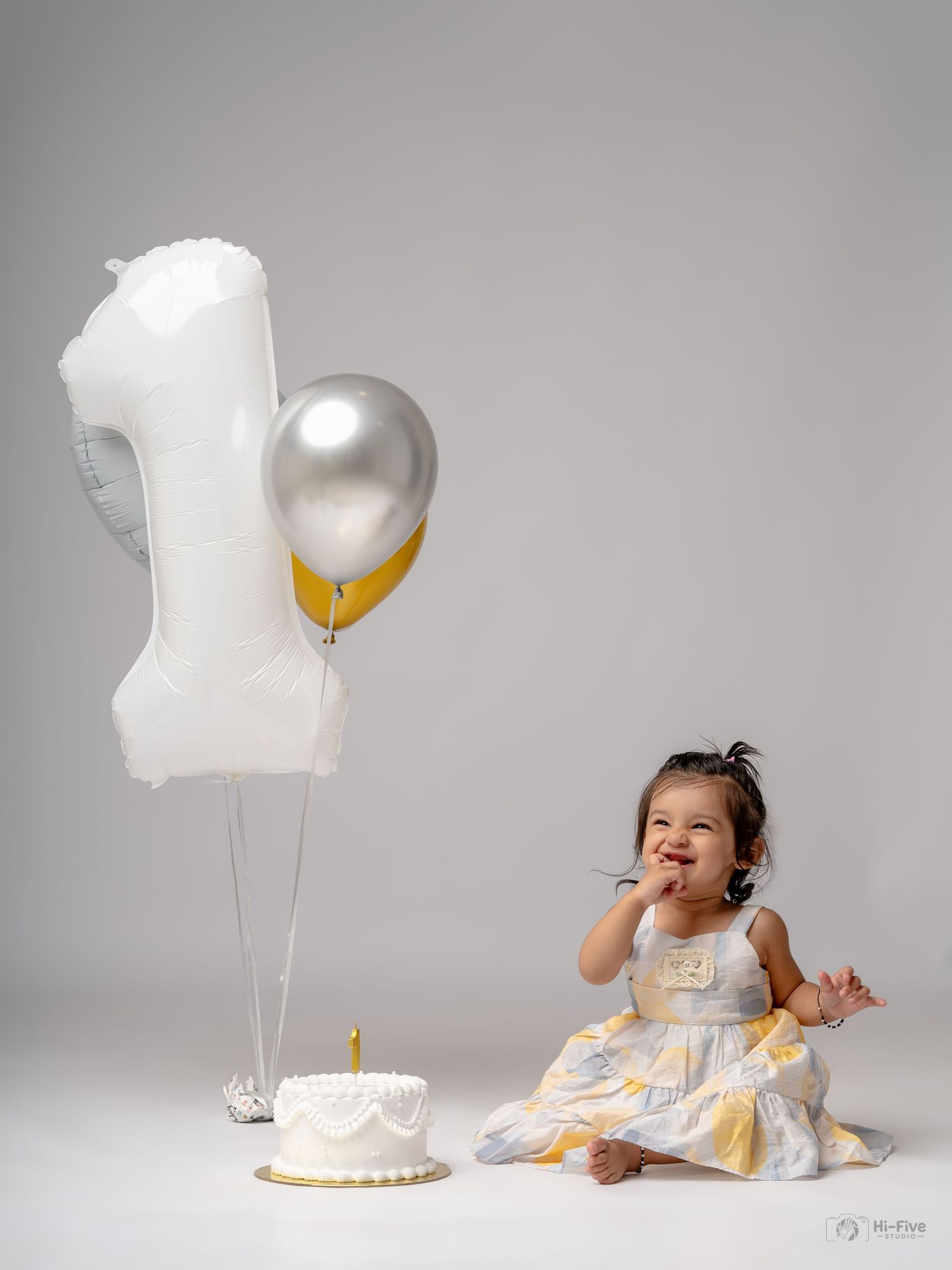 A baby girl in a floral dress sits beside a first birthday cake and number one balloon, smiling and tasting frosting.