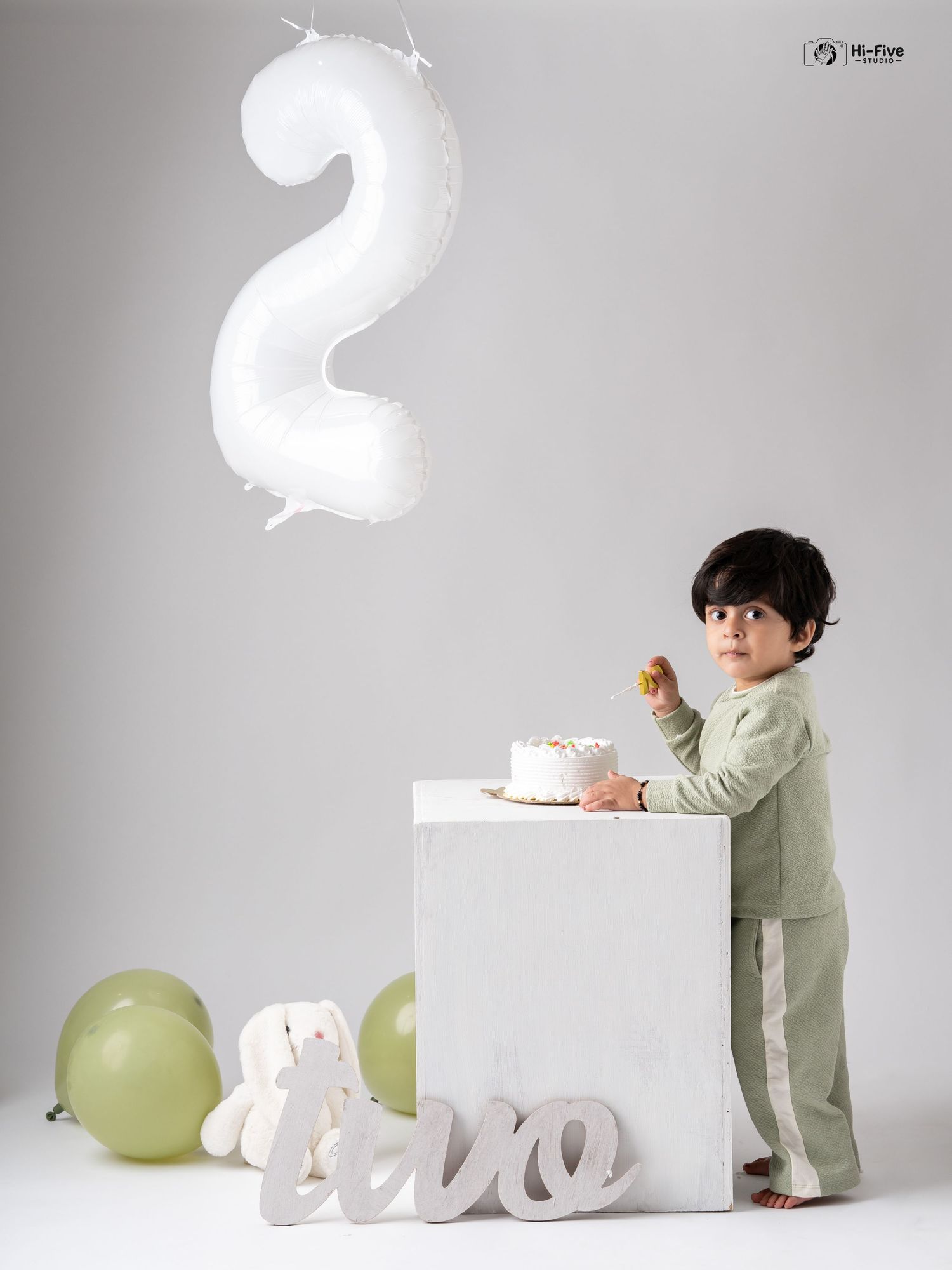 A toddler in green pajamas stands at a white cube touching a birthday cake, surrounded by balloons and a number 2 balloon.