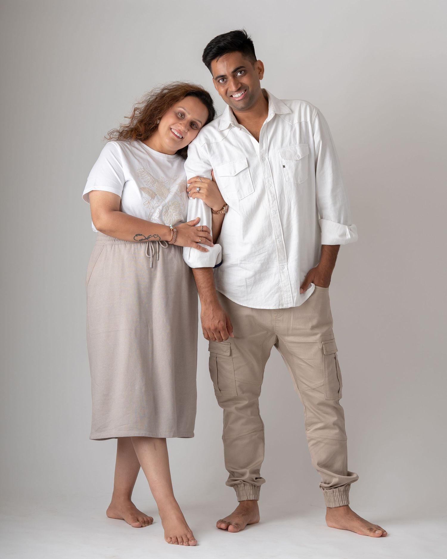 A smiling couple in coordinating white and beige outfits posing together barefoot in a studio portrait.