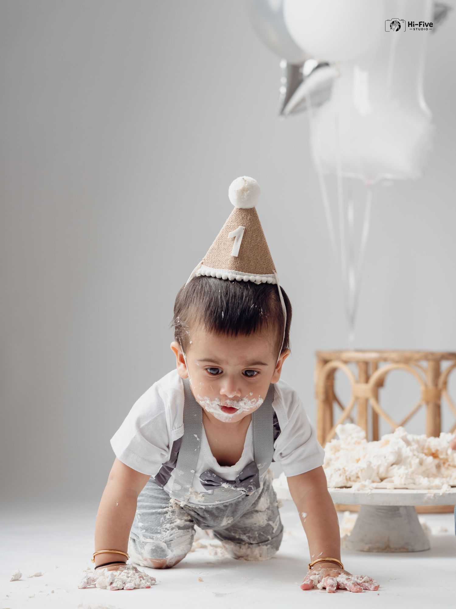 Toddler boy with party hat crawling with cake smash mess on hands during first birthday shoot.