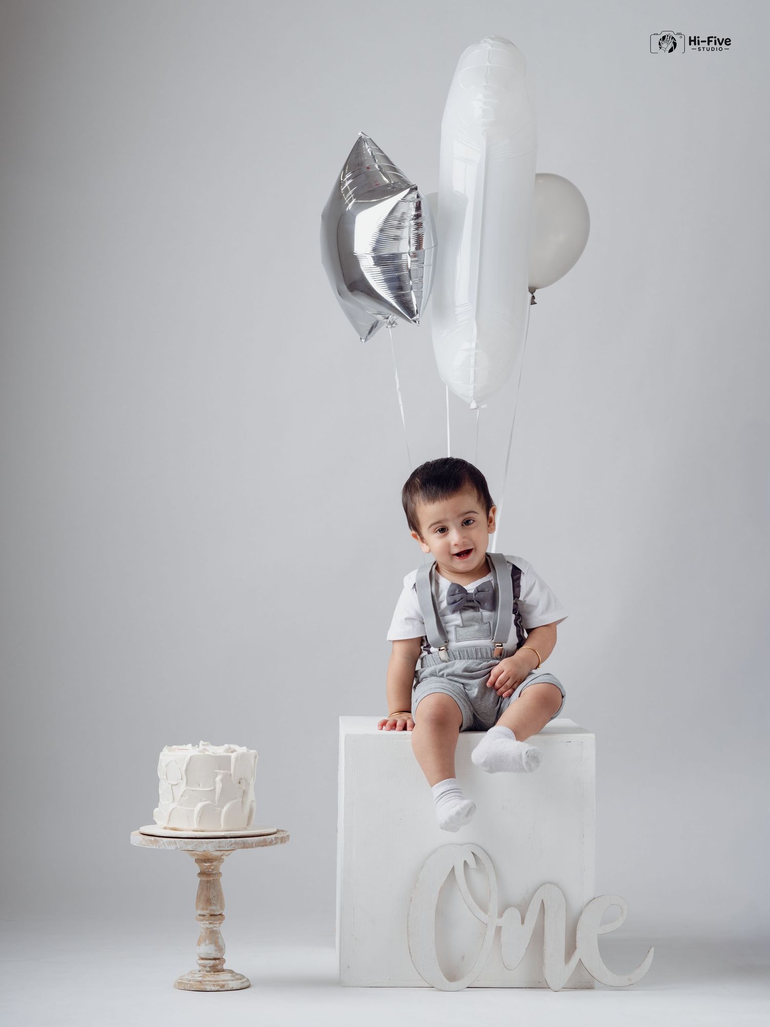 Happy toddler boy sitting on white cube with balloons and birthday cake for first birthday photo.