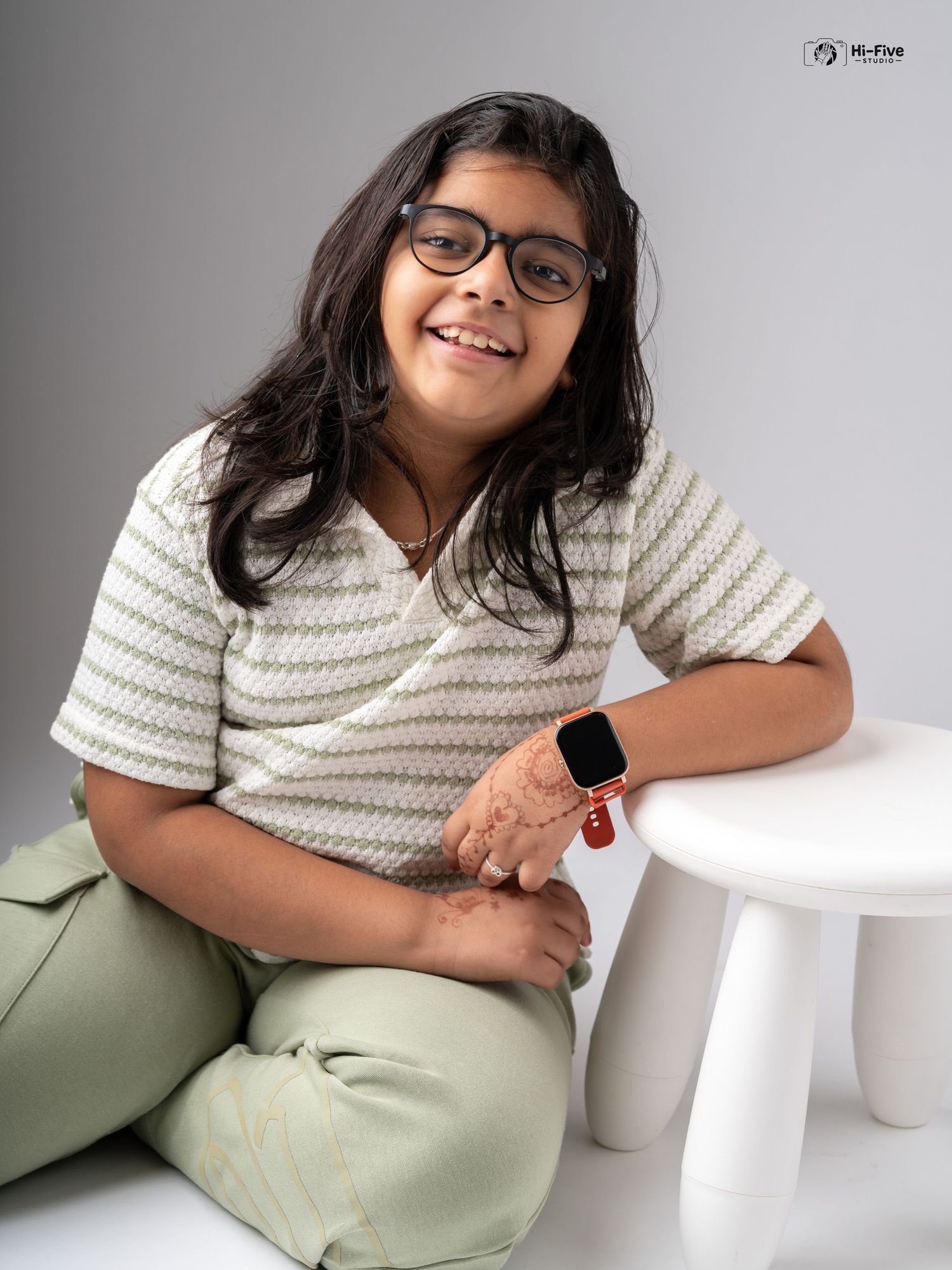 A cheerful girl with glasses sits cross-legged beside a white stool, wearing a striped top and green pants with a smartwatch.
