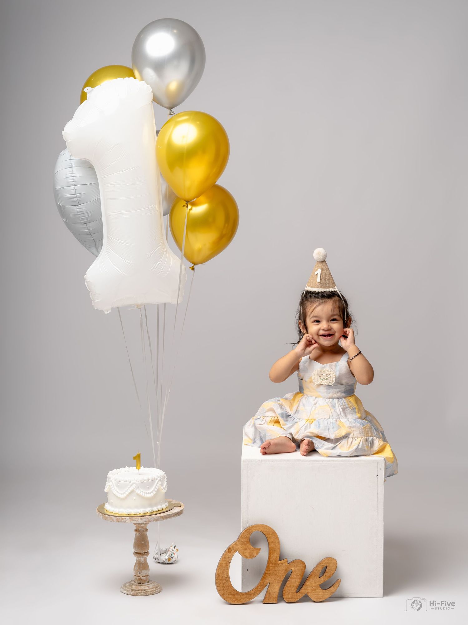 Baby girl in white gold dress celebrates first birthday with number one balloon bouquet and smash cake.
