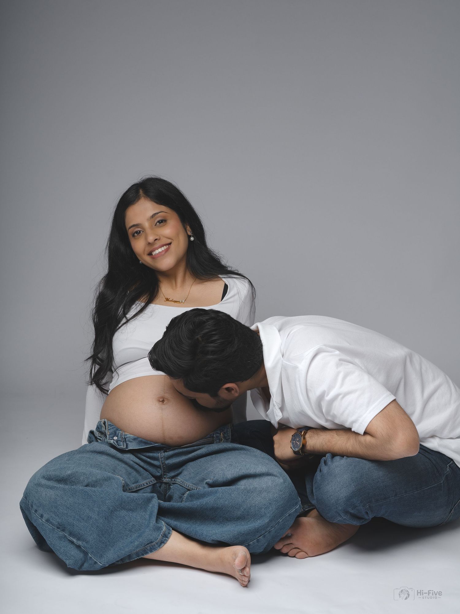 Smiling pregnant woman in white crop top while partner lovingly kisses her baby bump on floor.