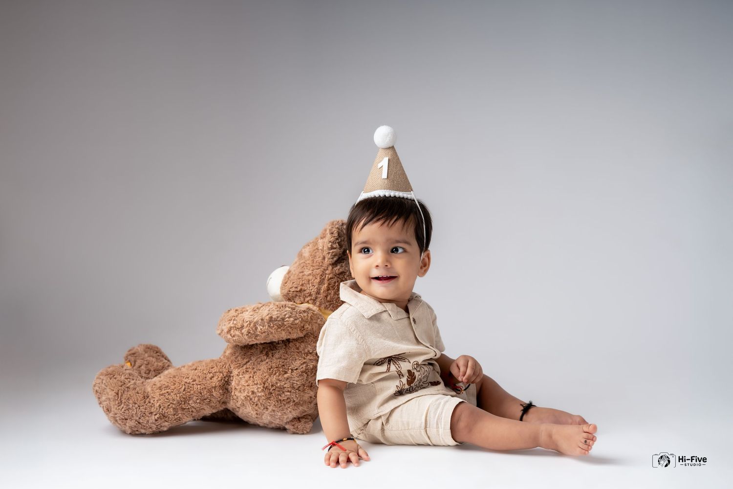 A baby boy in a birthday hat sits beside a large teddy bear in a professional studio photo.