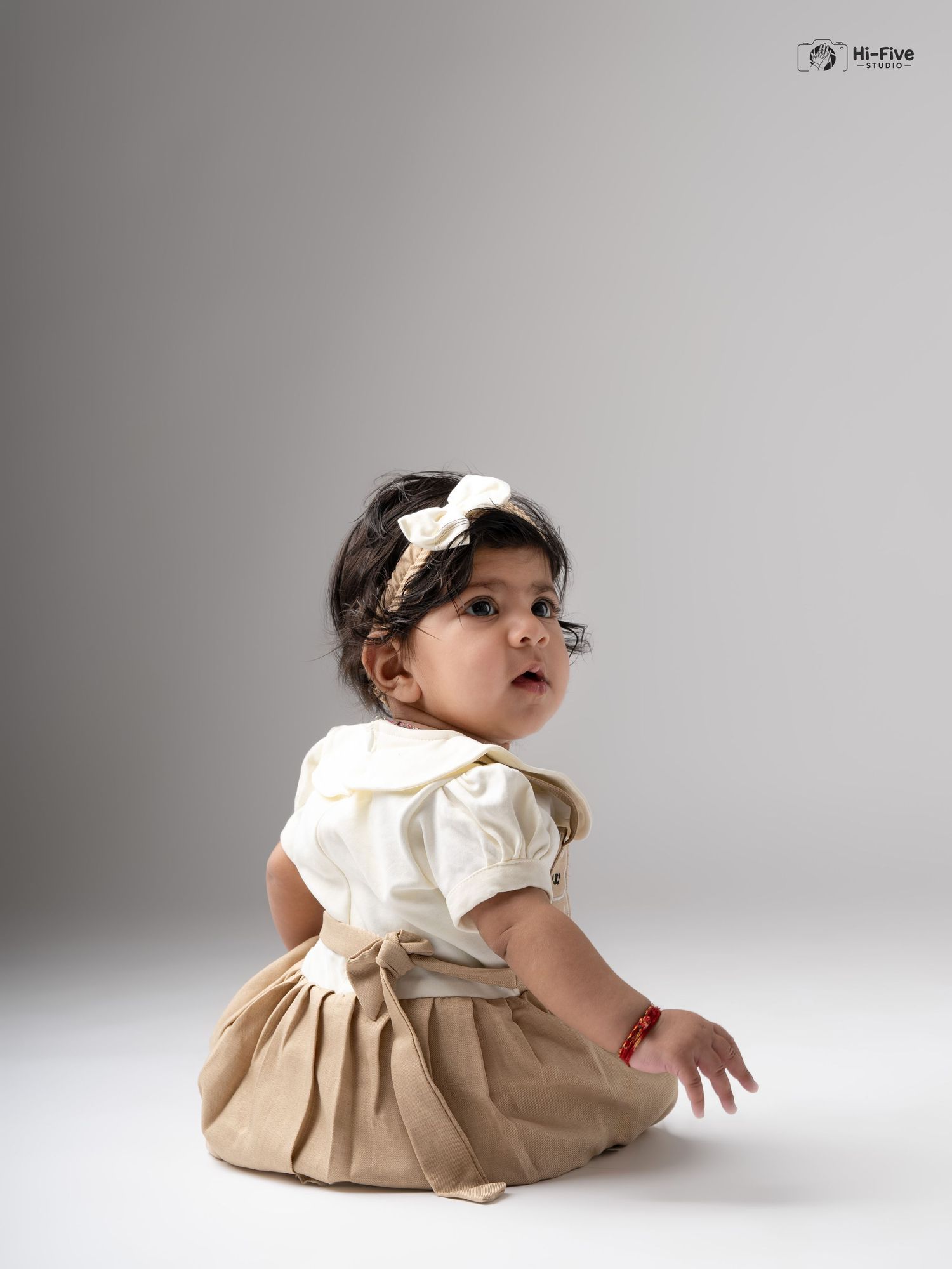 Adorable Indian baby girl in cream and tan outfit with bow headband sits in a professional studio photo.