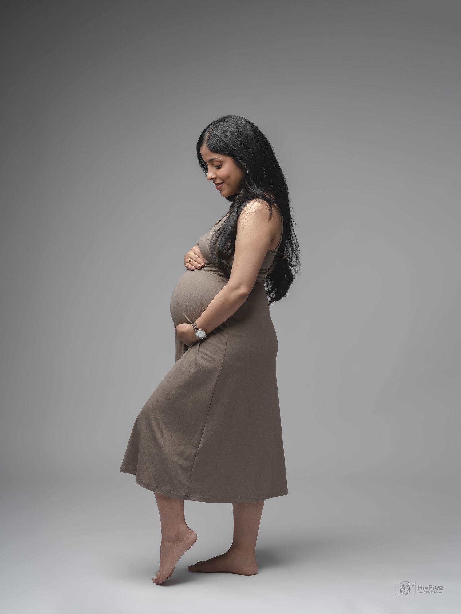 Pregnant woman in taupe midi dress tenderly holding baby bump in elegant side-profile studio portrait.