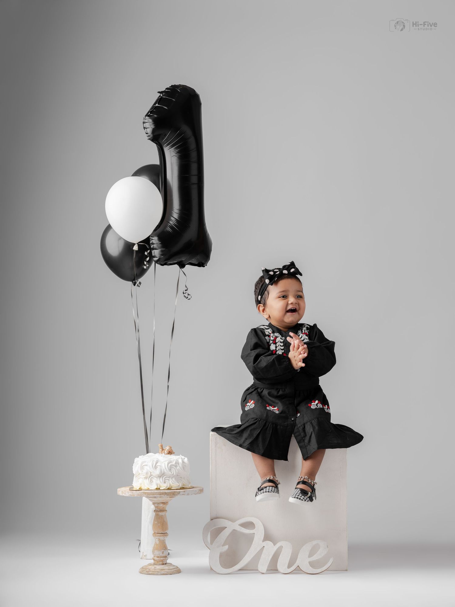 Baby girl in black dress celebrates first birthday with balloons, cake, and One sign in studio photo.