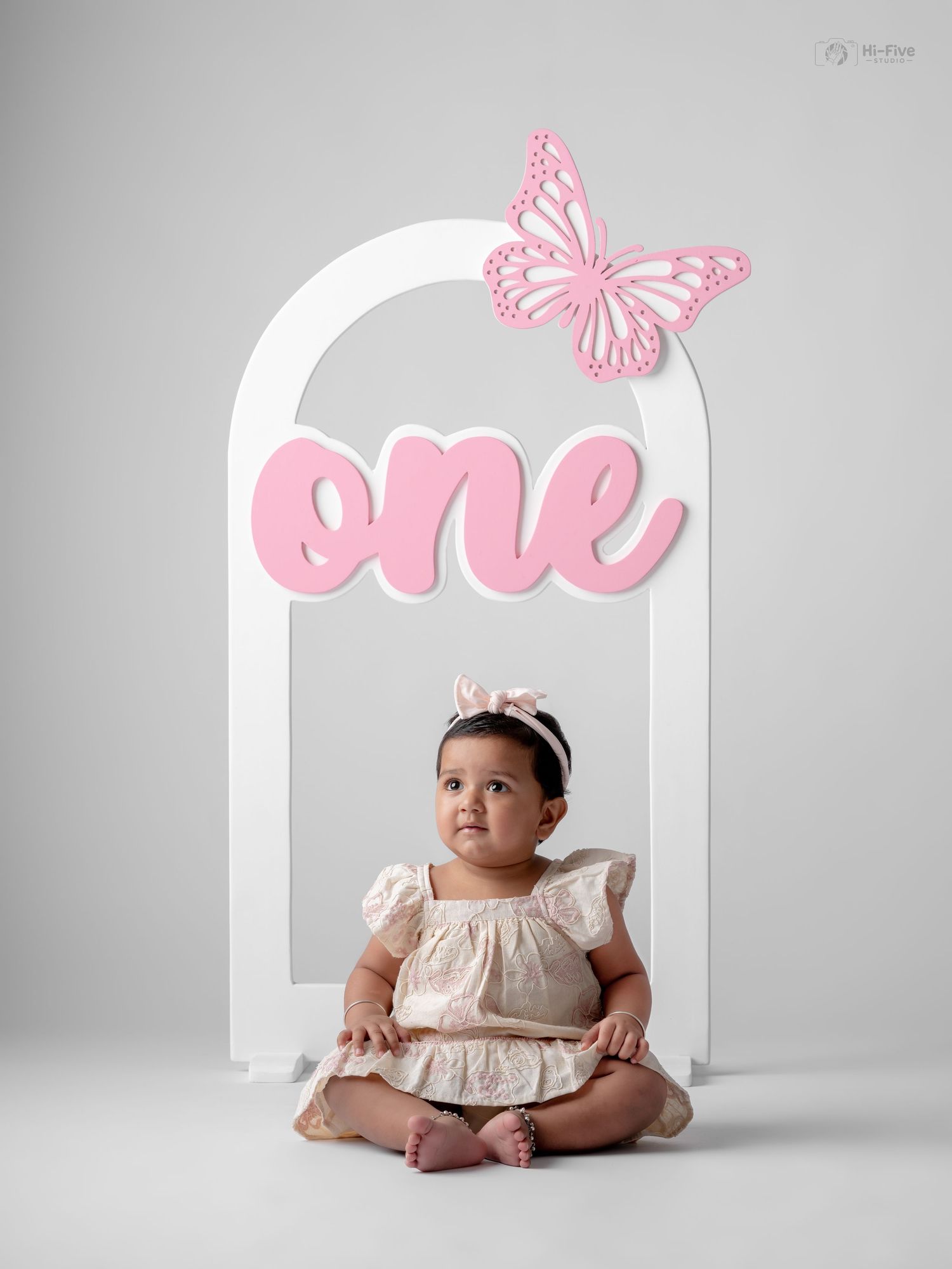 Baby girl in cream dress sits before white arch with pink butterfly and \