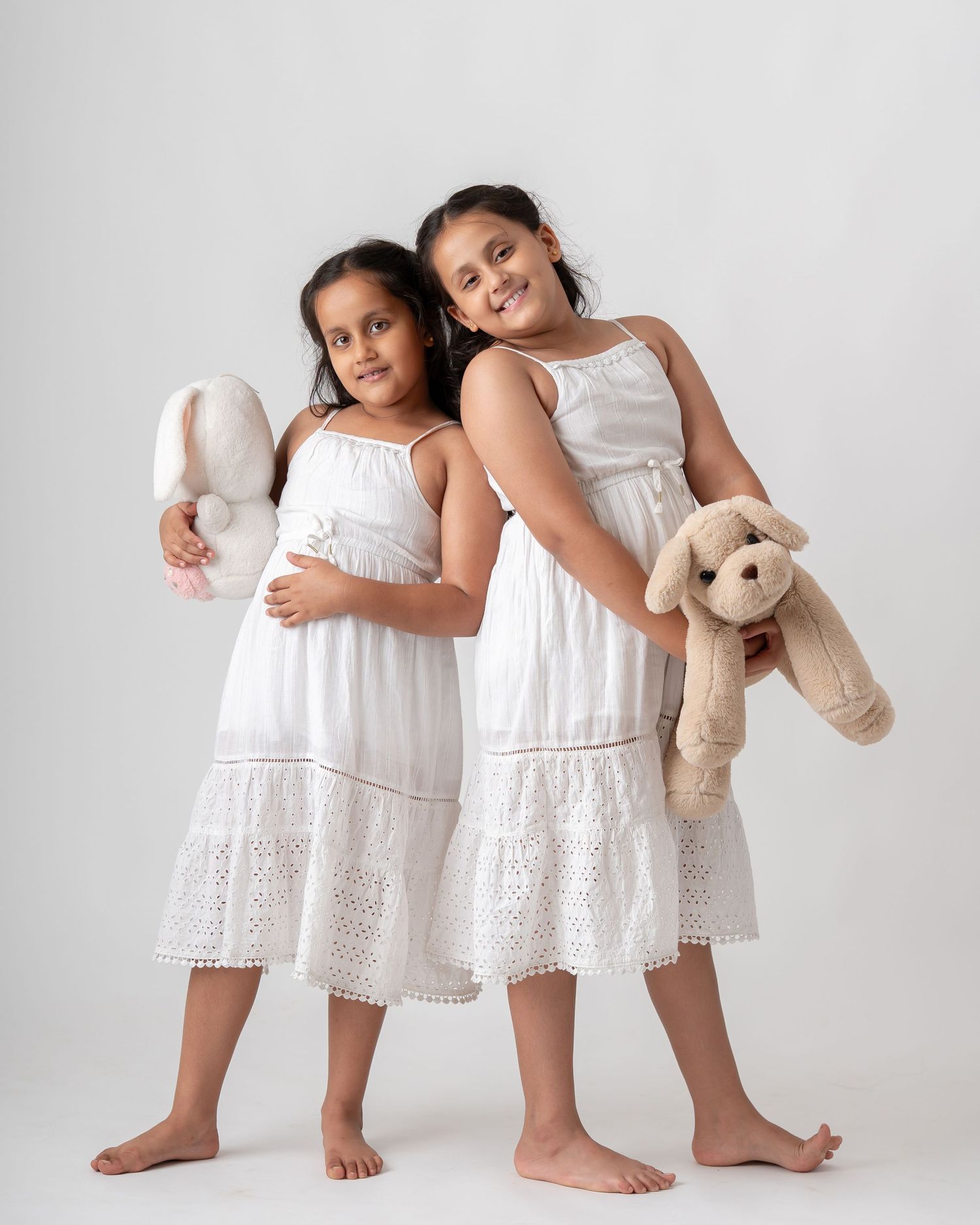 Two young girls in white eyelet dresses holding stuffed animal toys in a studio portrait.