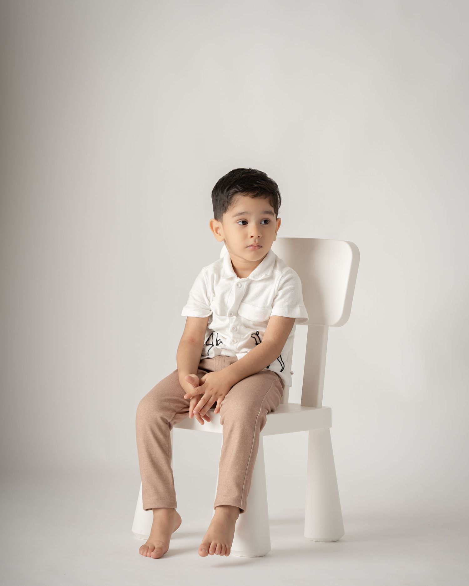 Young boy in white shirt and beige pants sitting on a white chair in a studio portrait.