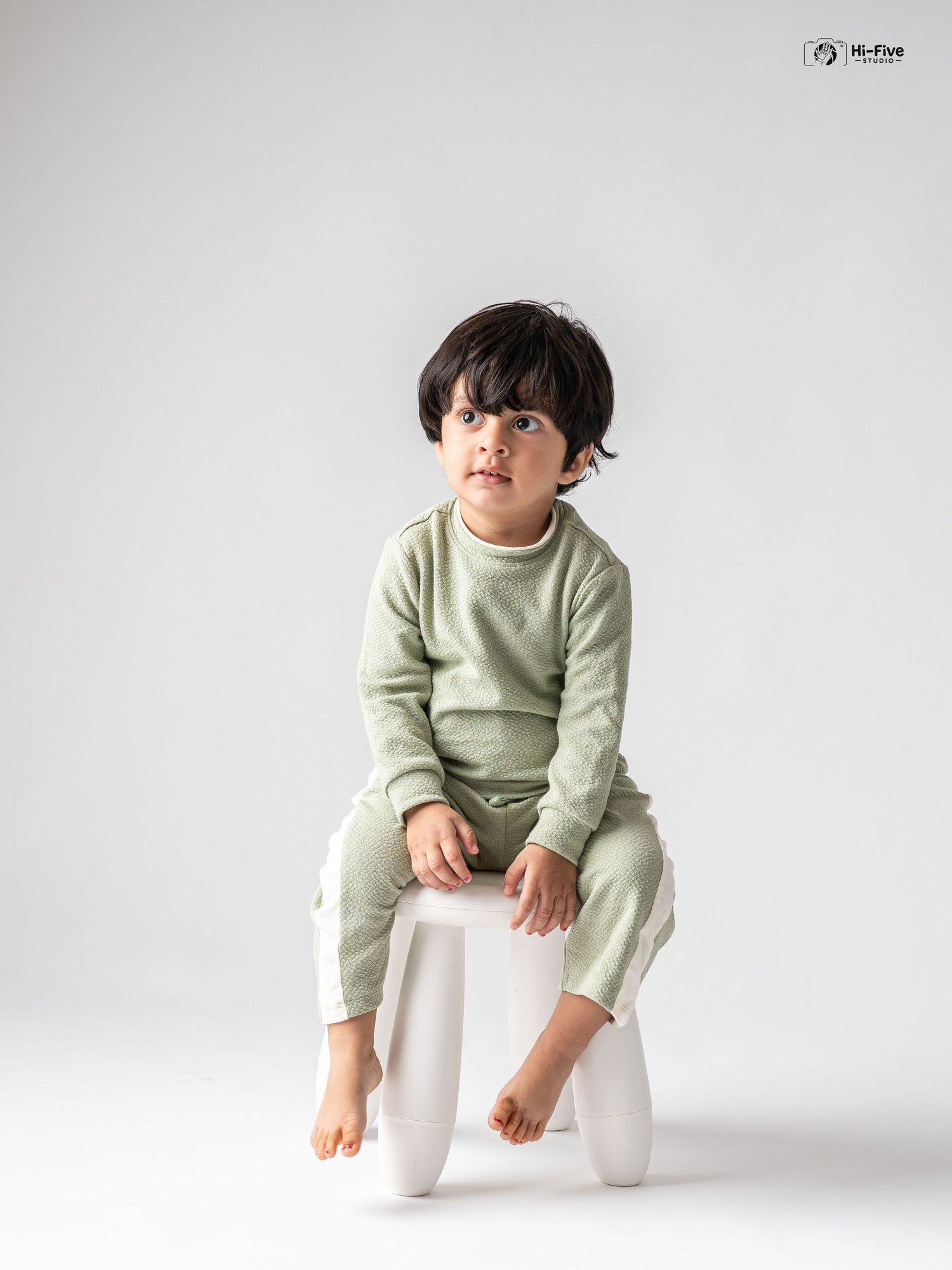 Young toddler boy in sage green matching sweatsuit sitting on white stool in studio photo.