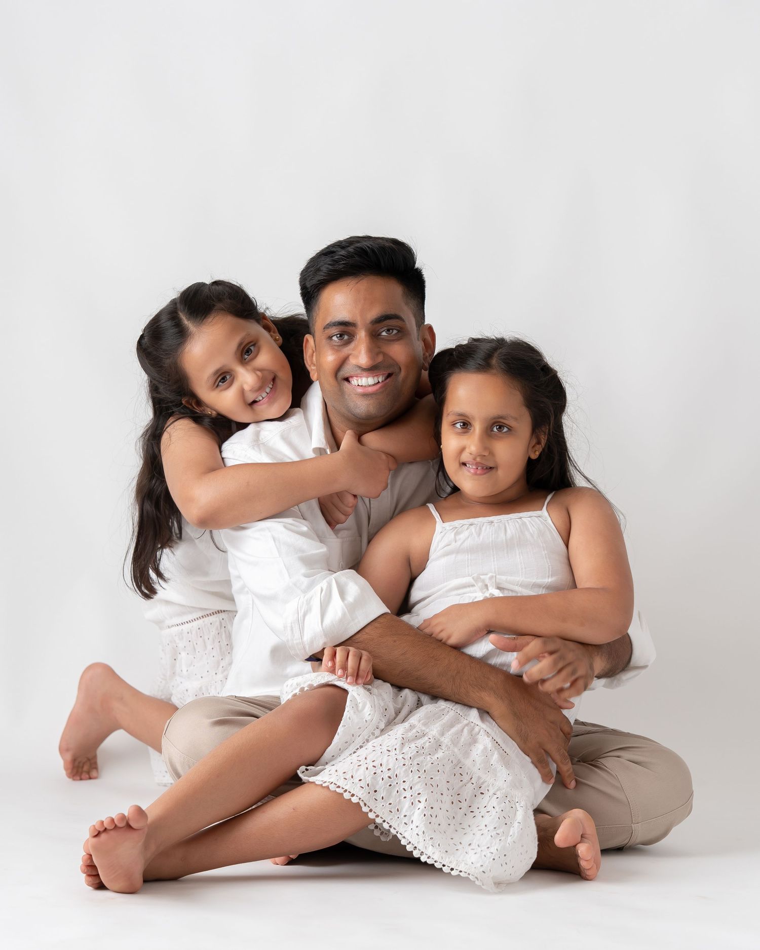 Man sitting cross-legged with two young girls in white dresses hugging him in a studio portrait.