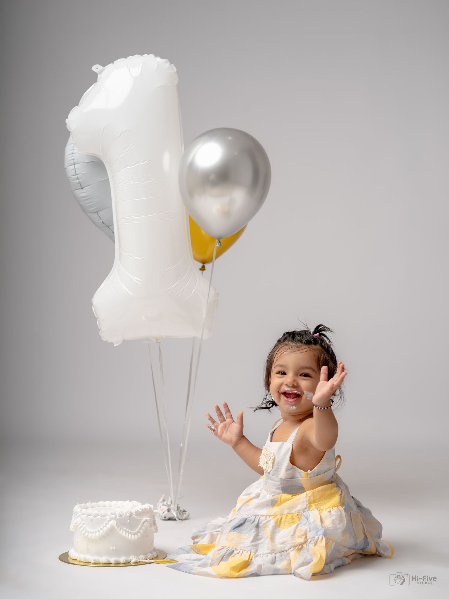 Smiling baby girl in white dress celebrates 1st birthday with number one balloon, silver and gold balloons, and cake.