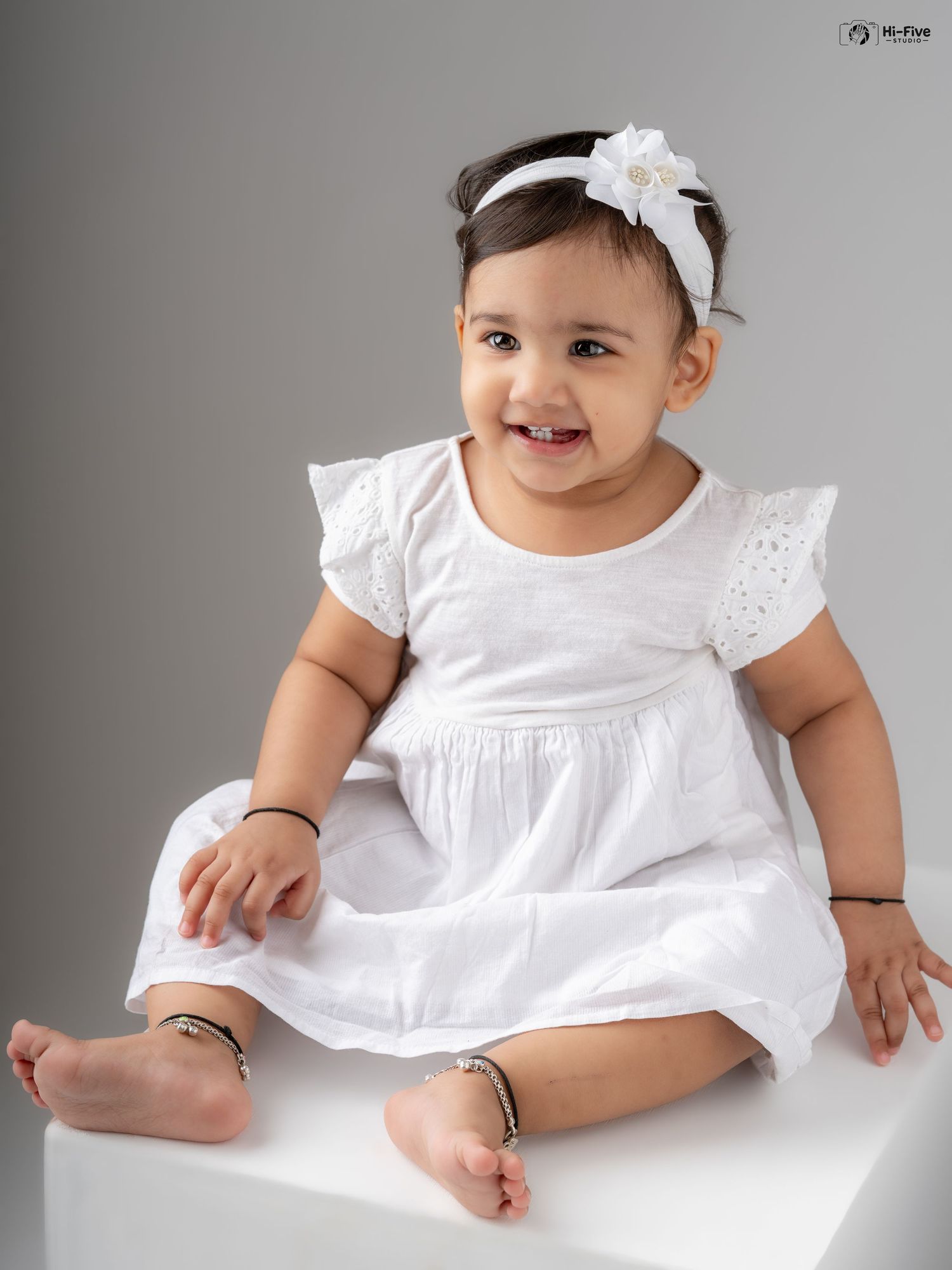 Cheerful baby girl in white dress and bow headband sitting barefoot on white cube smiling.