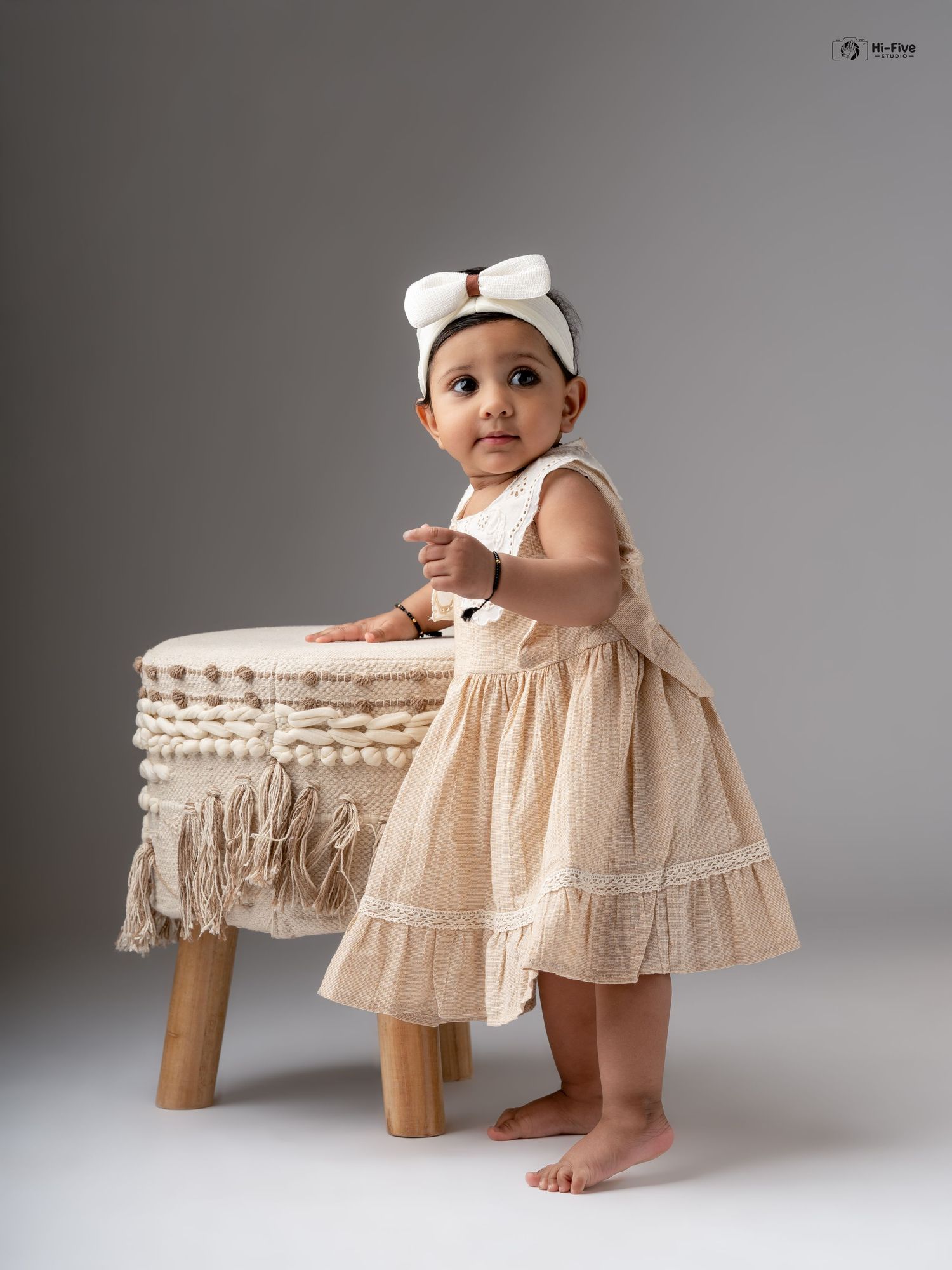 Adorable baby girl in beige linen dress with white bow headband stands beside boho stool in studio.