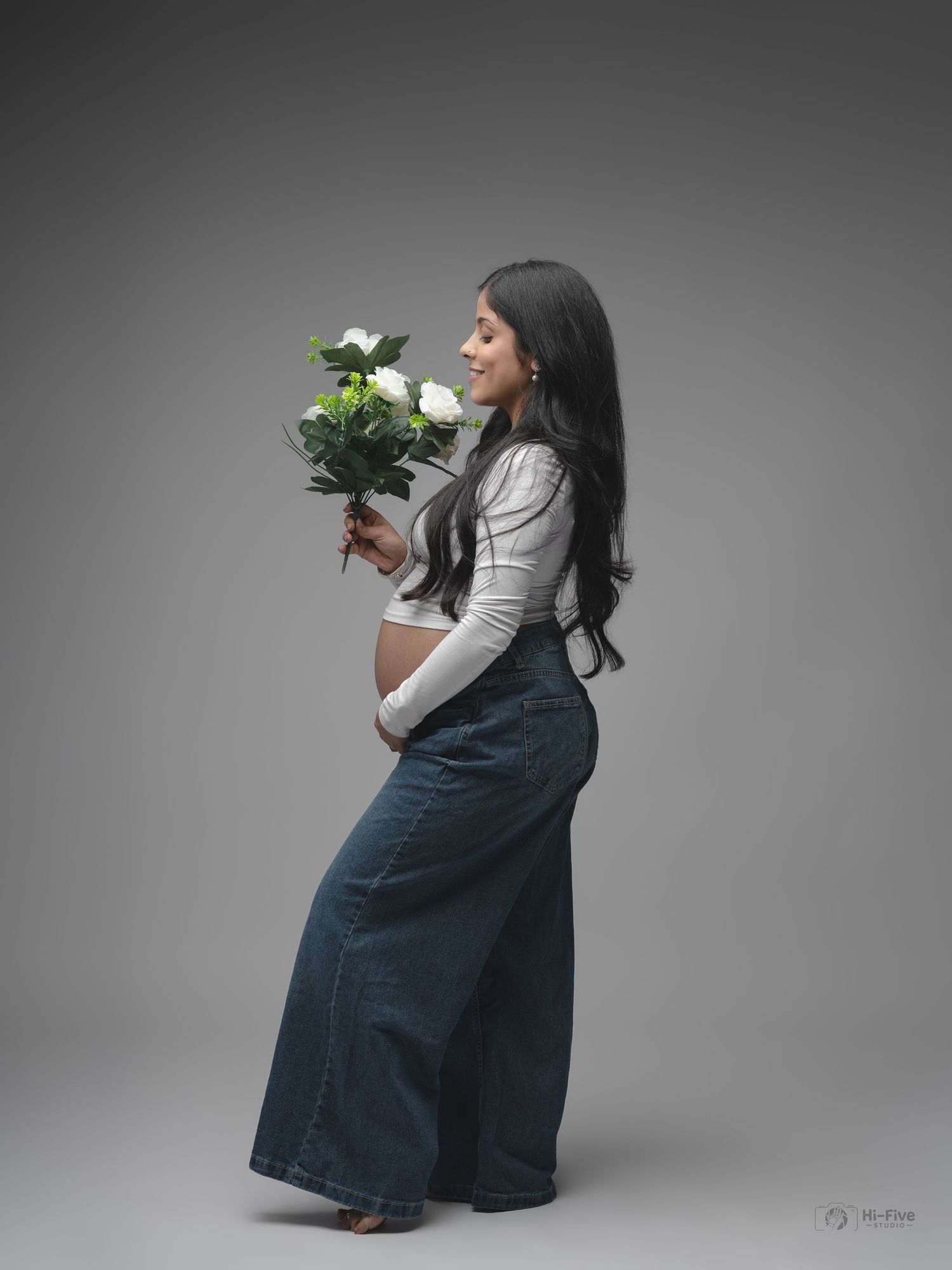 Pregnant woman in denim skirt holding white flowers bouquet during elegant maternity photoshoot.