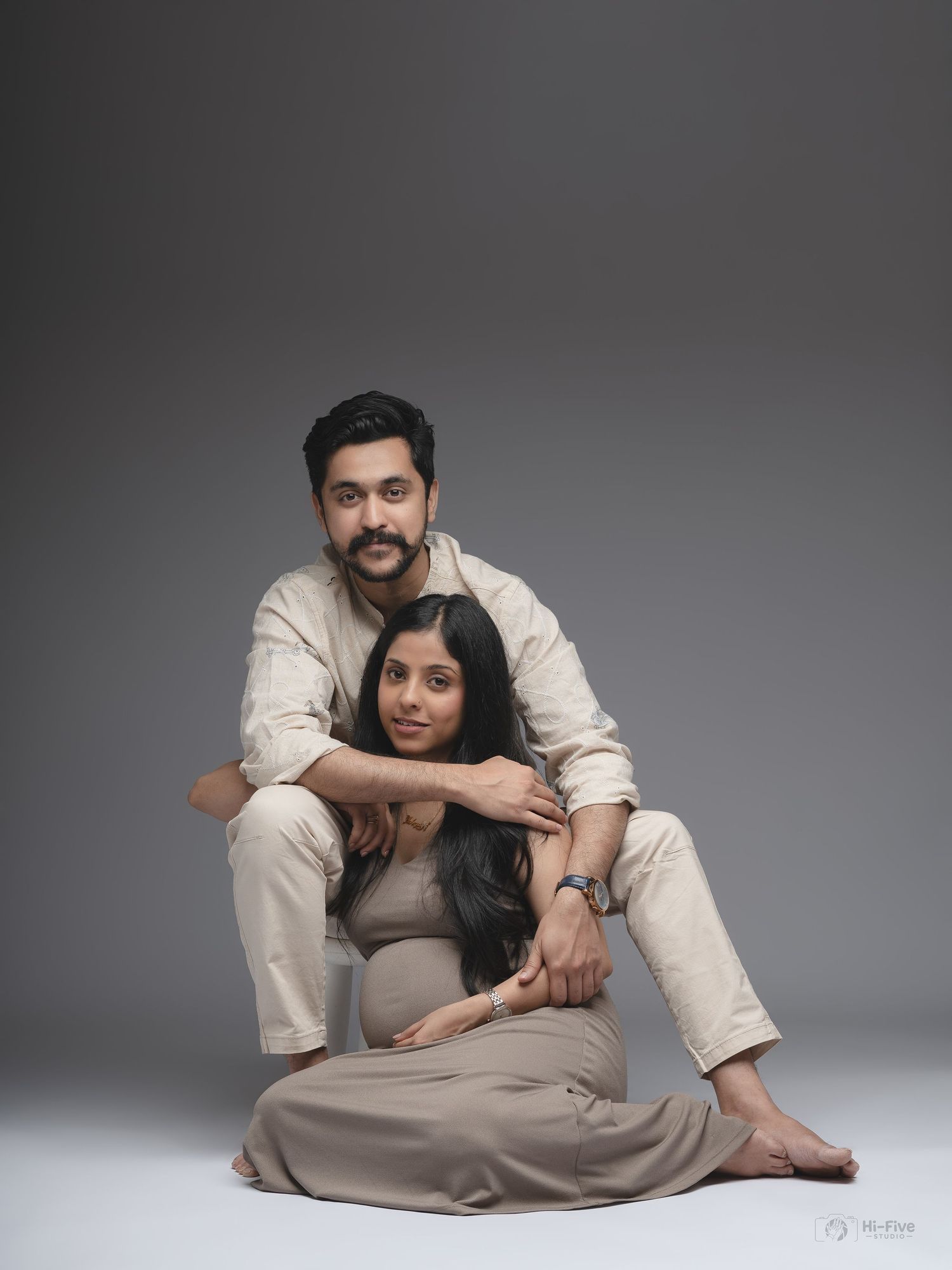 Couple posing for maternity photoshoot in matching beige outfits against neutral gray studio backdrop.