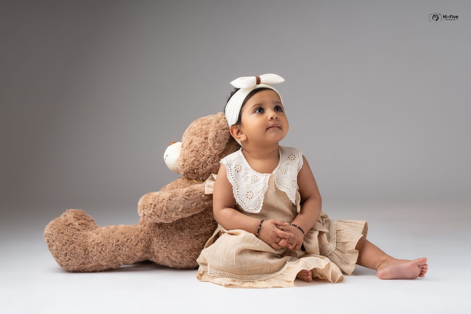 Sweet baby girl in tan romper sits with large teddy bear, wearing white bow headband in studio.