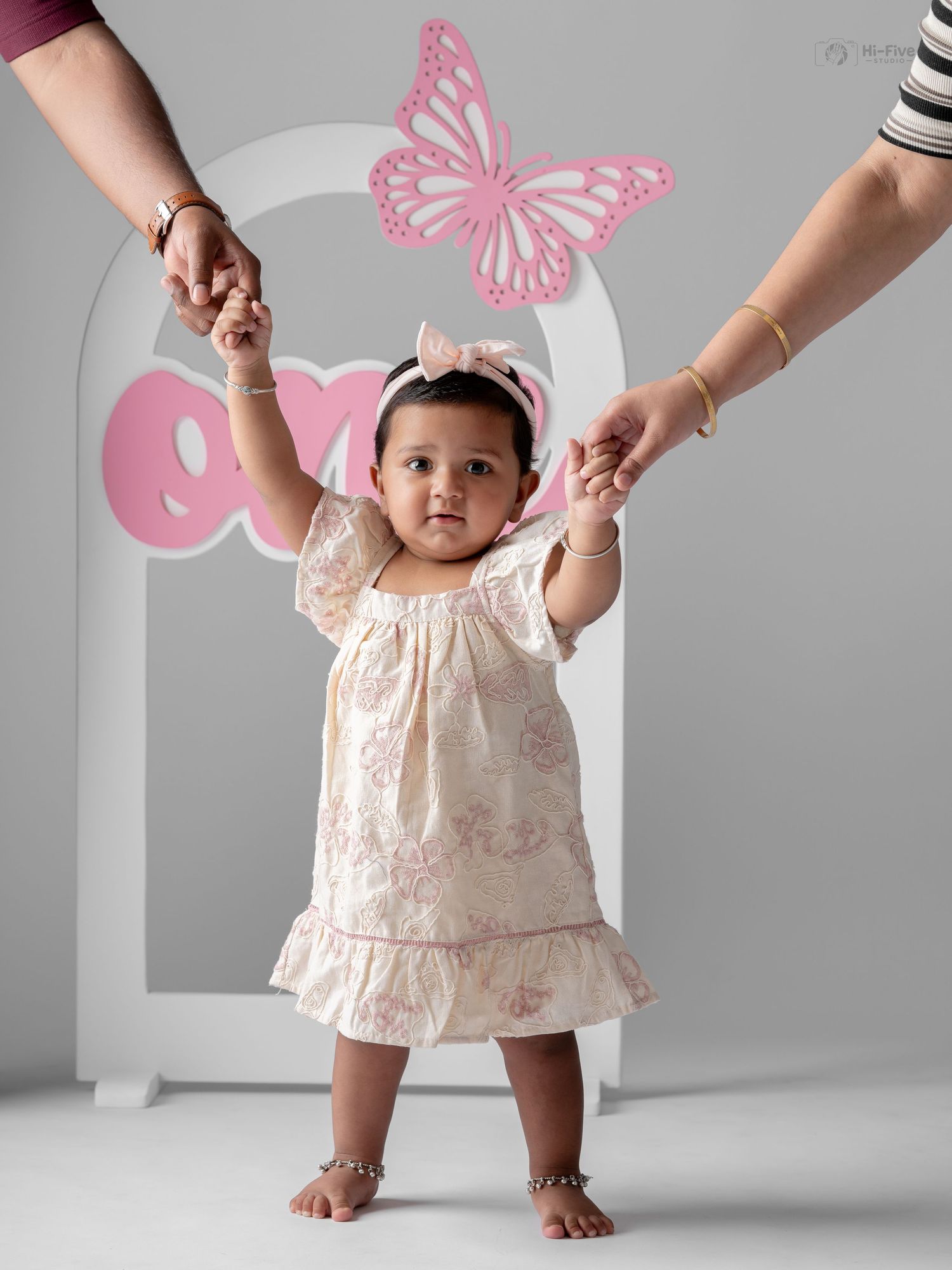 Baby girl in floral dress stands holding adults hands with pink butterfly decor backdrop.