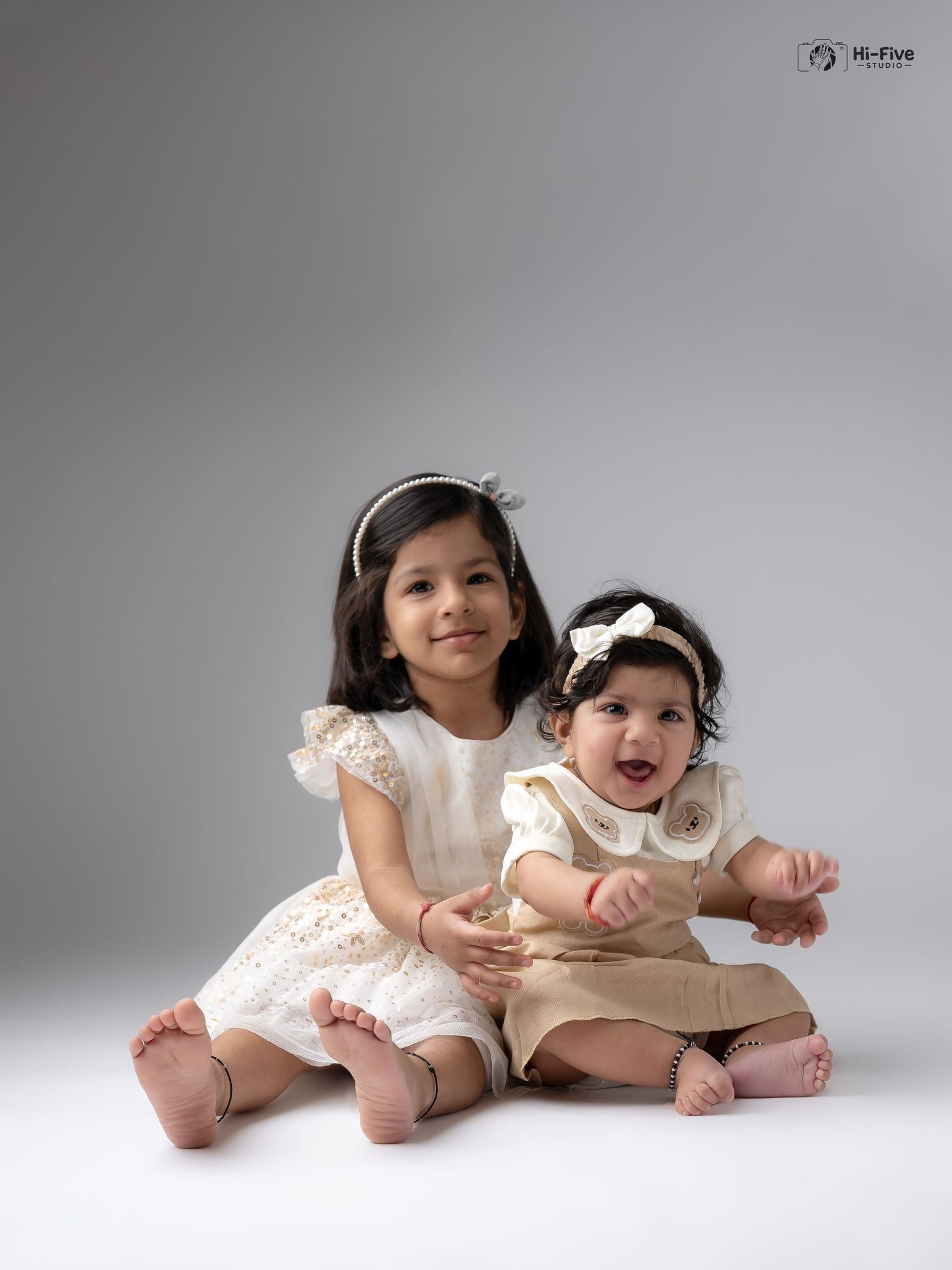 Two adorable Indian sisters in cream and gold dresses with headbands pose together in a professional studio photo.
