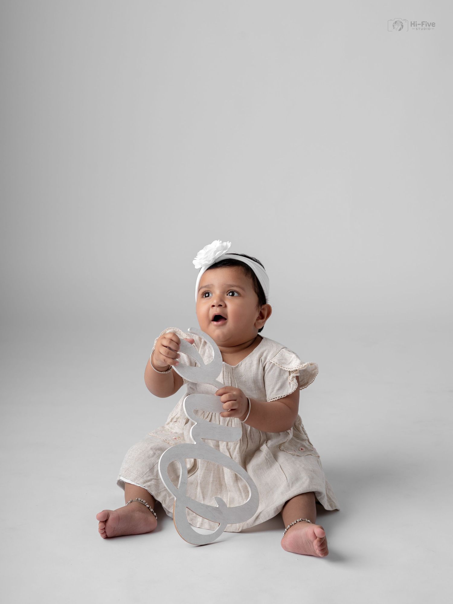 A cute baby girl in a white dress and headband sits on a studio floor, holding a decorative letter while looking up.