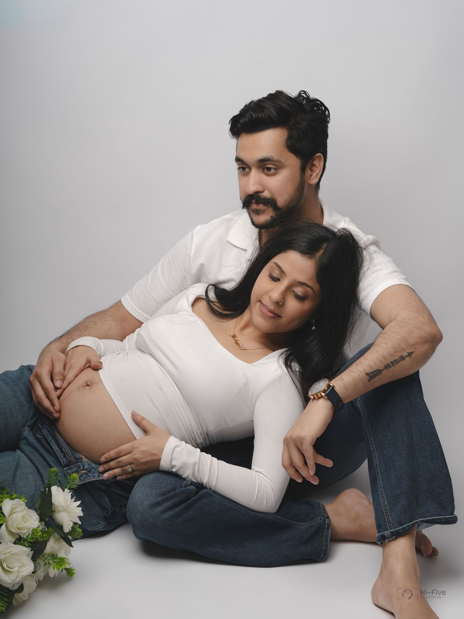 Couple maternity portrait with man embracing pregnant woman leaning on him with white flowers nearby.