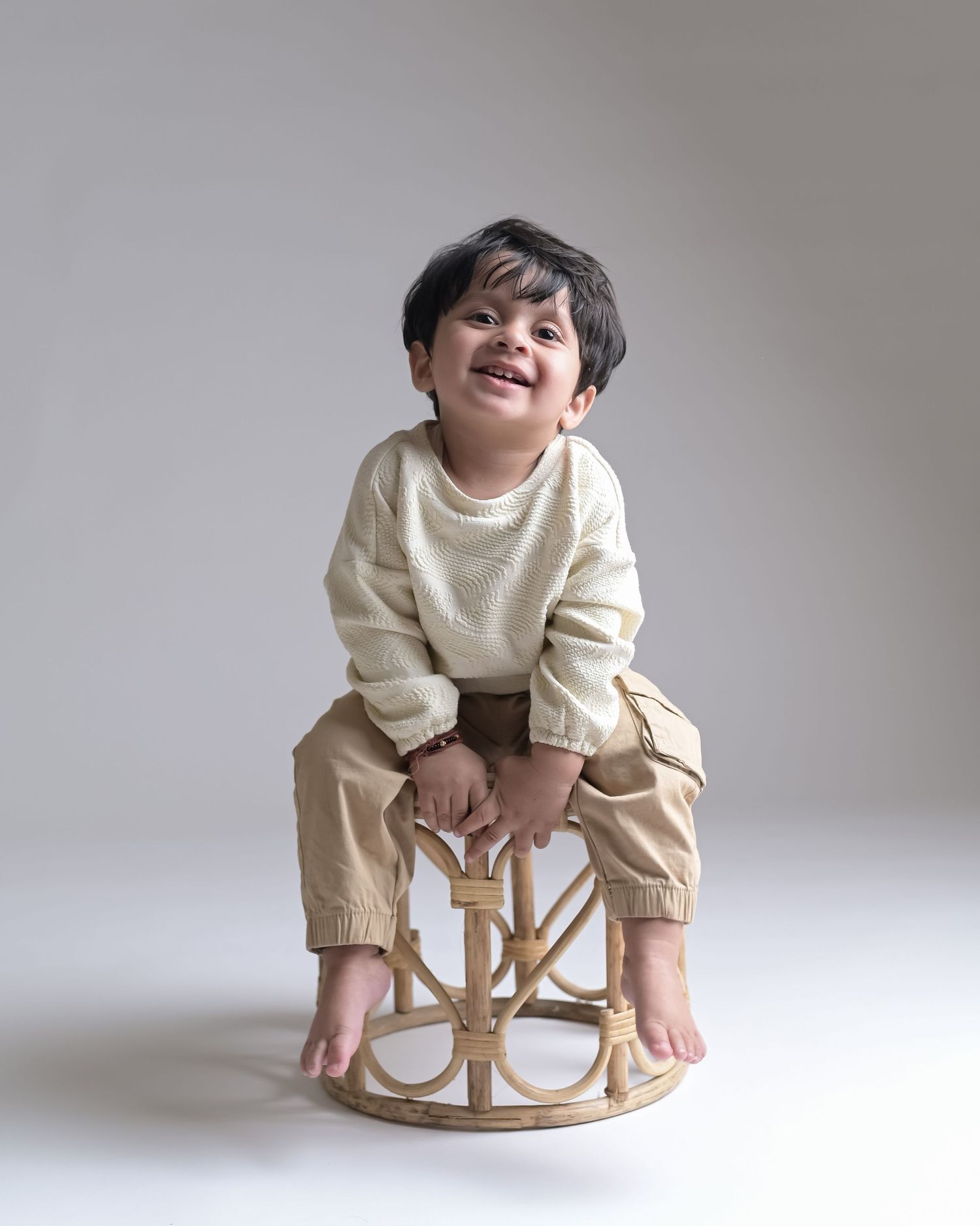 Happy toddler boy laughing on rattan stool in cream knit sweater and khaki pants.