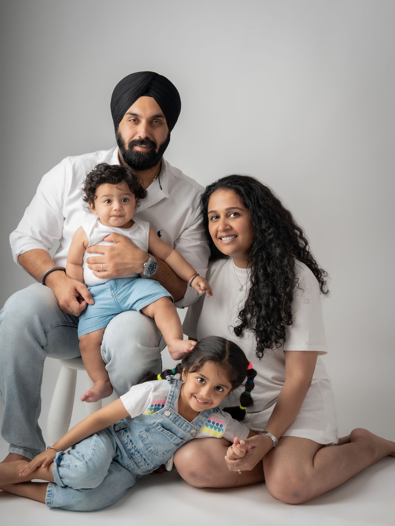 Sikh family of four with turban-wearing father holding baby and young daughter smiling.