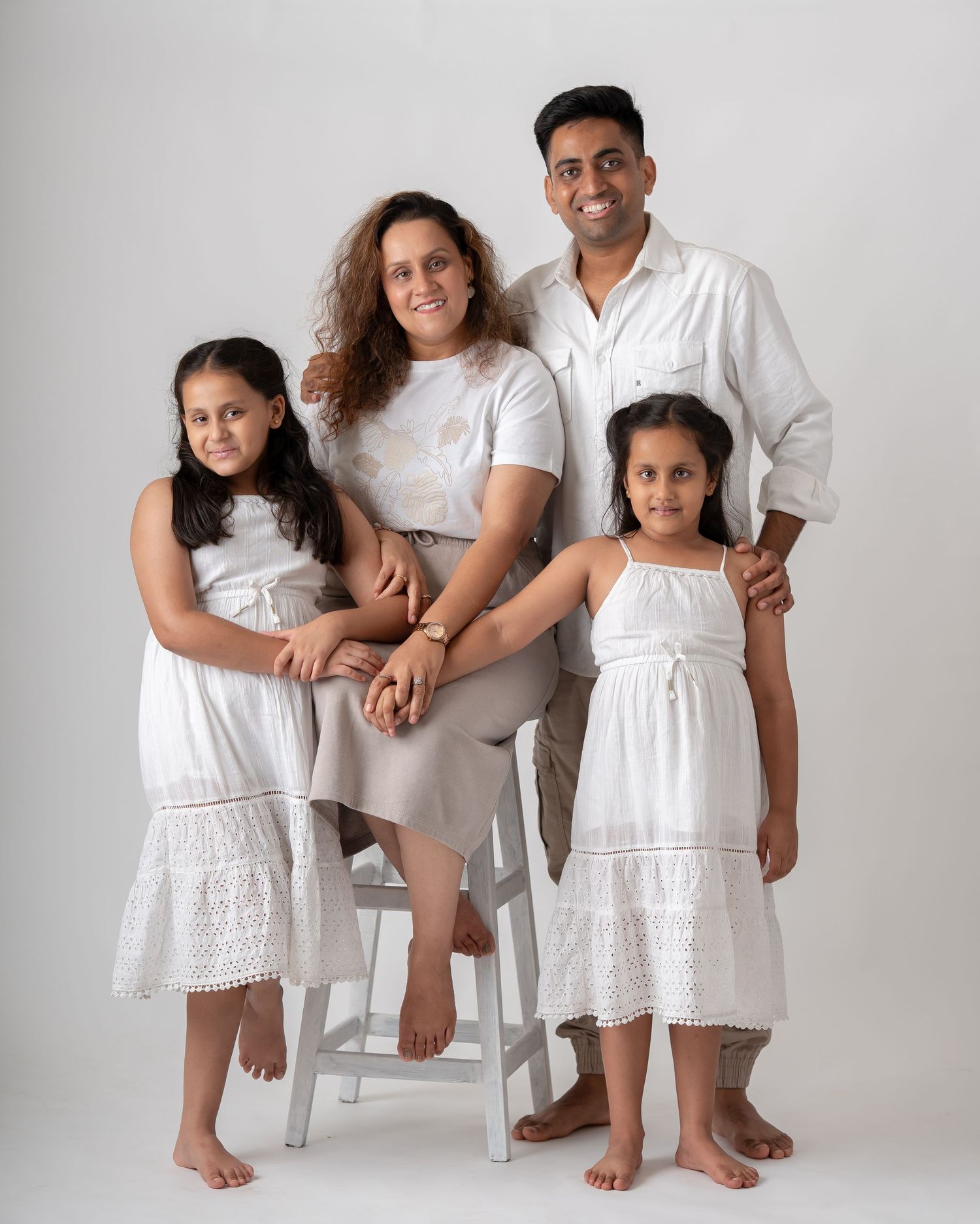 A family of four in white outfits poses together in a bright studio, with two young girls and their smiling parents.