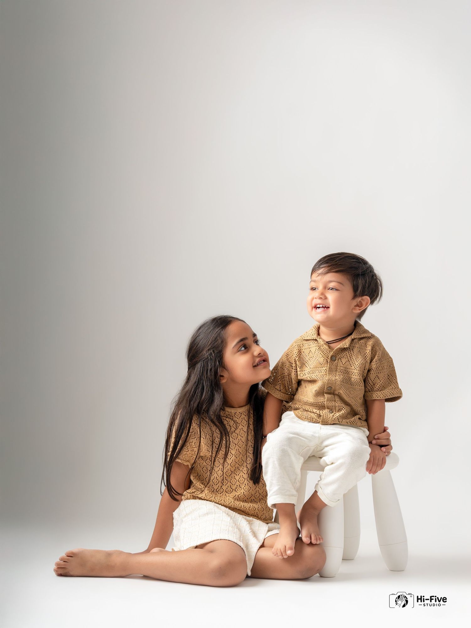 Two young siblings in matching gold tops and white shorts sitting together laughing in studio.